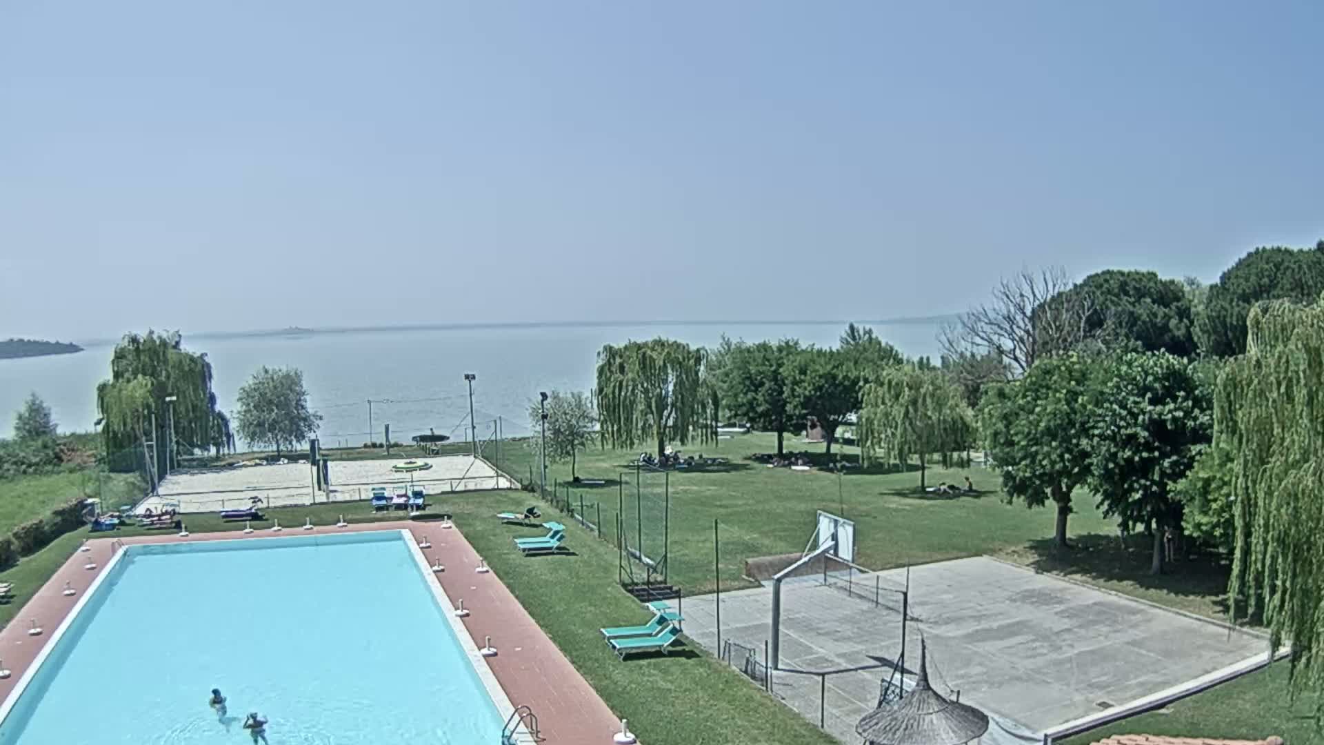 An outdoor swimming pool and recreational area with a basketball court and volleyball court are situated next to a calm lake on a sunny day.