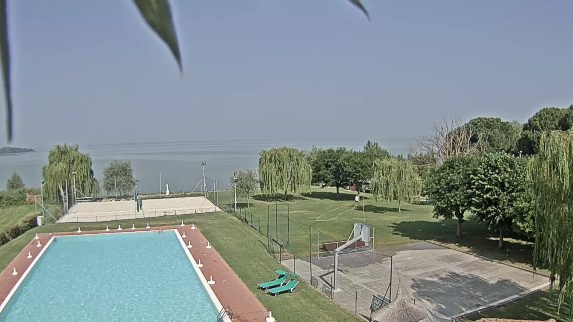 A swimming pool, volleyball court, and basketball court are situated on a grassy area overlooking a calm lake under a clear sky.