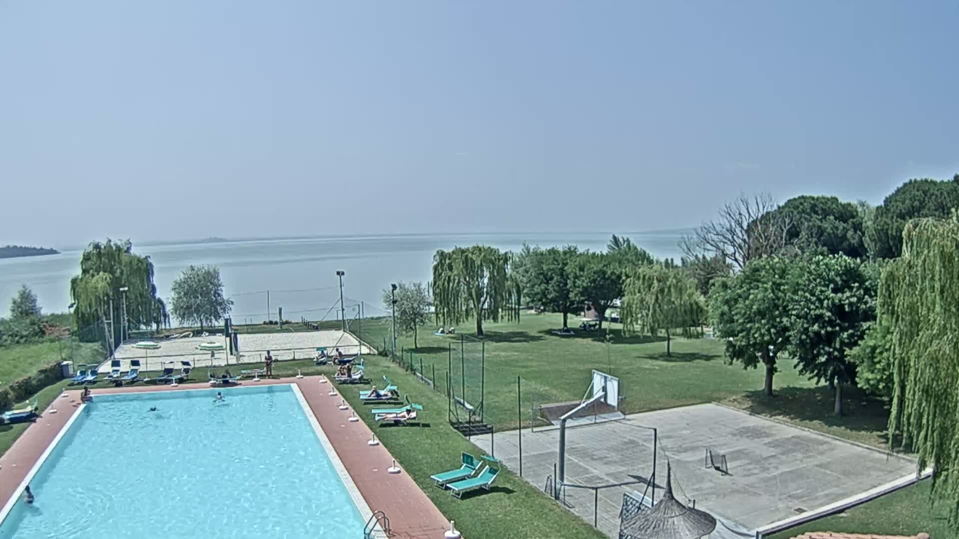 A swimming pool and a basketball court are situated on a grassy area overlooking a calm lake under a clear sky.