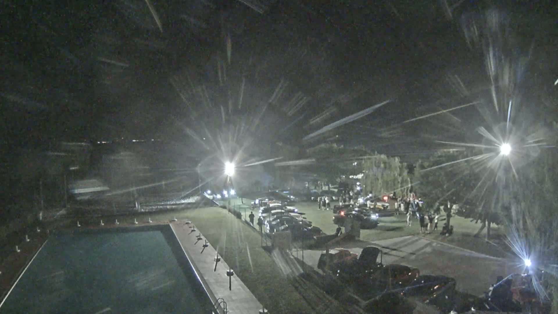 A nighttime aerial view shows a swimming pool, a parking lot filled with cars and people, and bright lights causing lens flare.