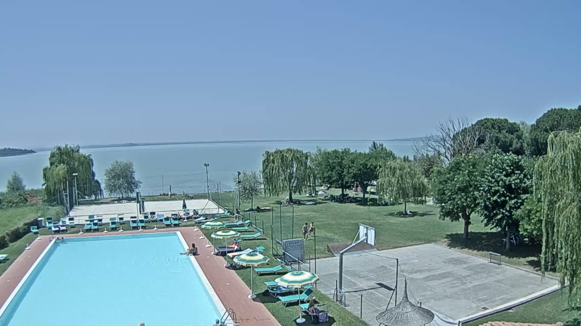 A swimming pool with lounge chairs and umbrellas sits beside a basketball court and a grassy area, all overlooking a calm lake under a clear blue sky.