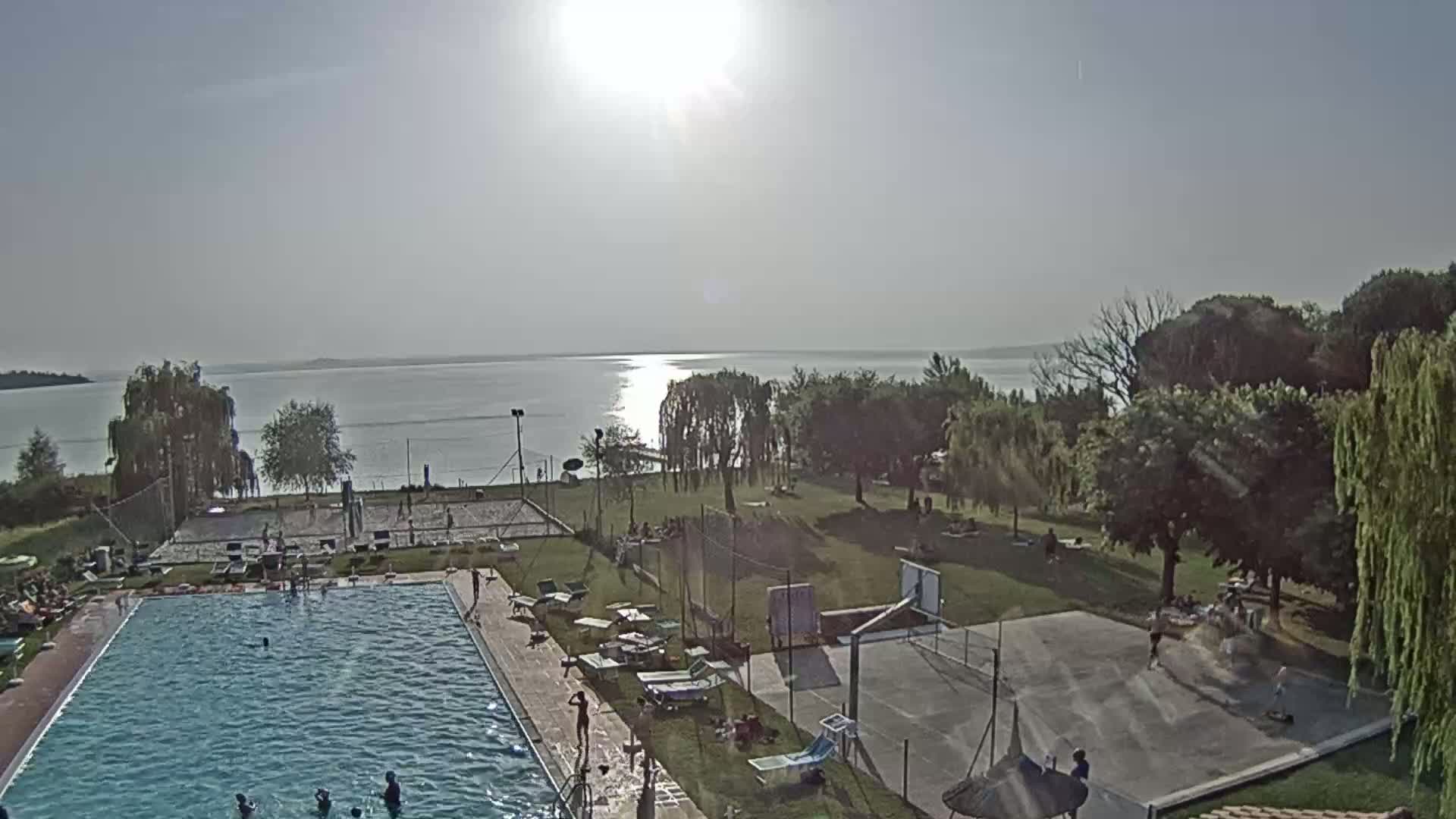 A sunny day overlooking a lake shows a swimming pool, volleyball courts, a basketball court, and people enjoying the recreational area.