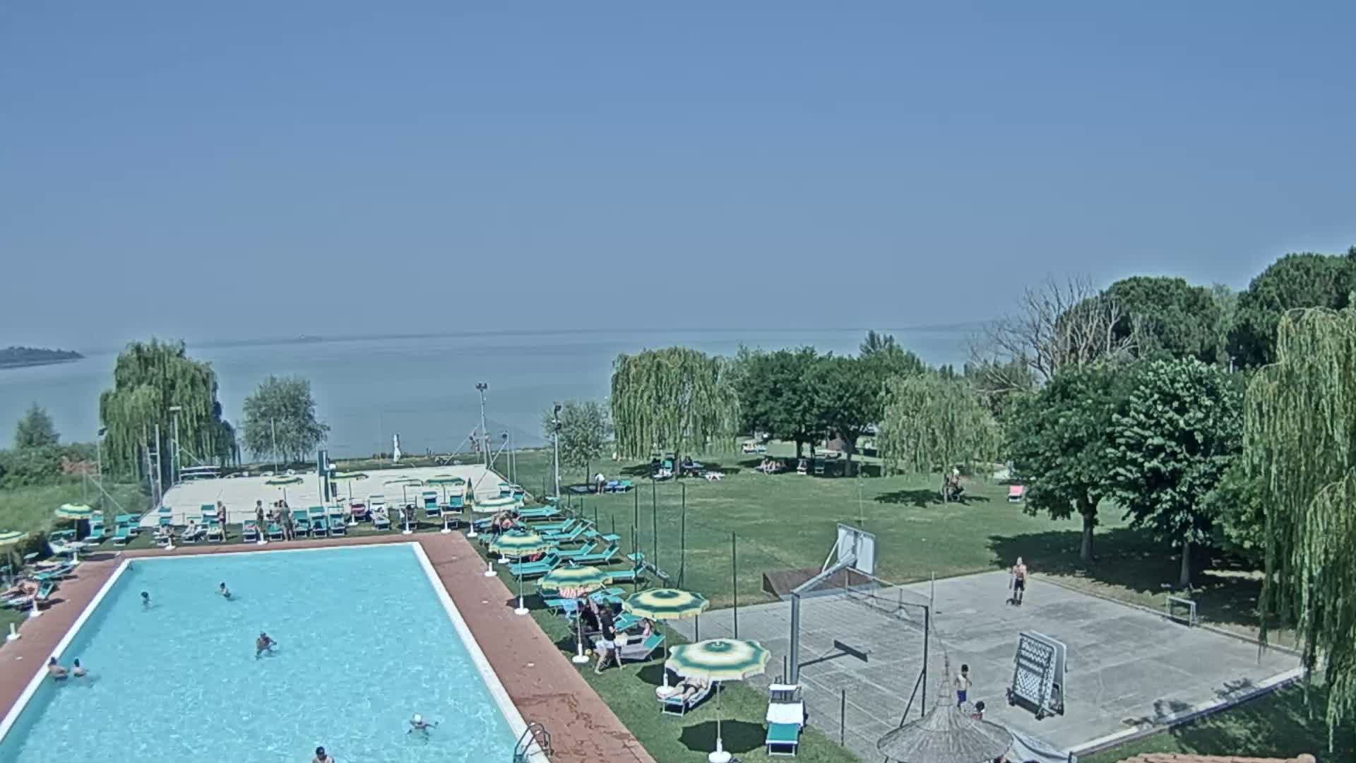 A swimming pool with people swimming and sunbathing is situated near a grassy area with a basketball court, all overlooking a calm lake on a clear, sunny day.