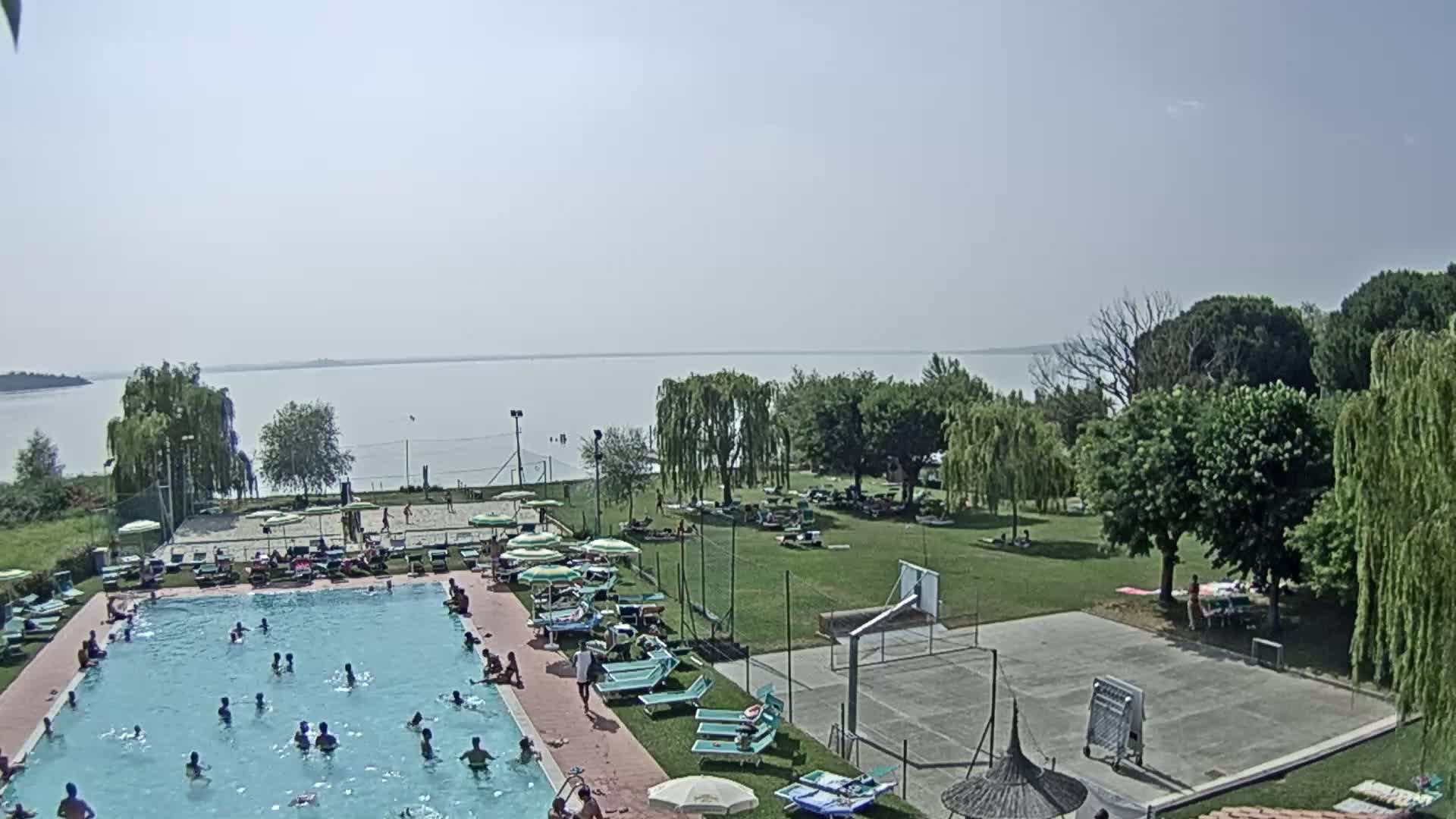 A swimming pool with many people in it is surrounded by a grassy area with trees, a volleyball court, a basketball court, and lounge chairs, all overlooking a calm lake on a sunny day.