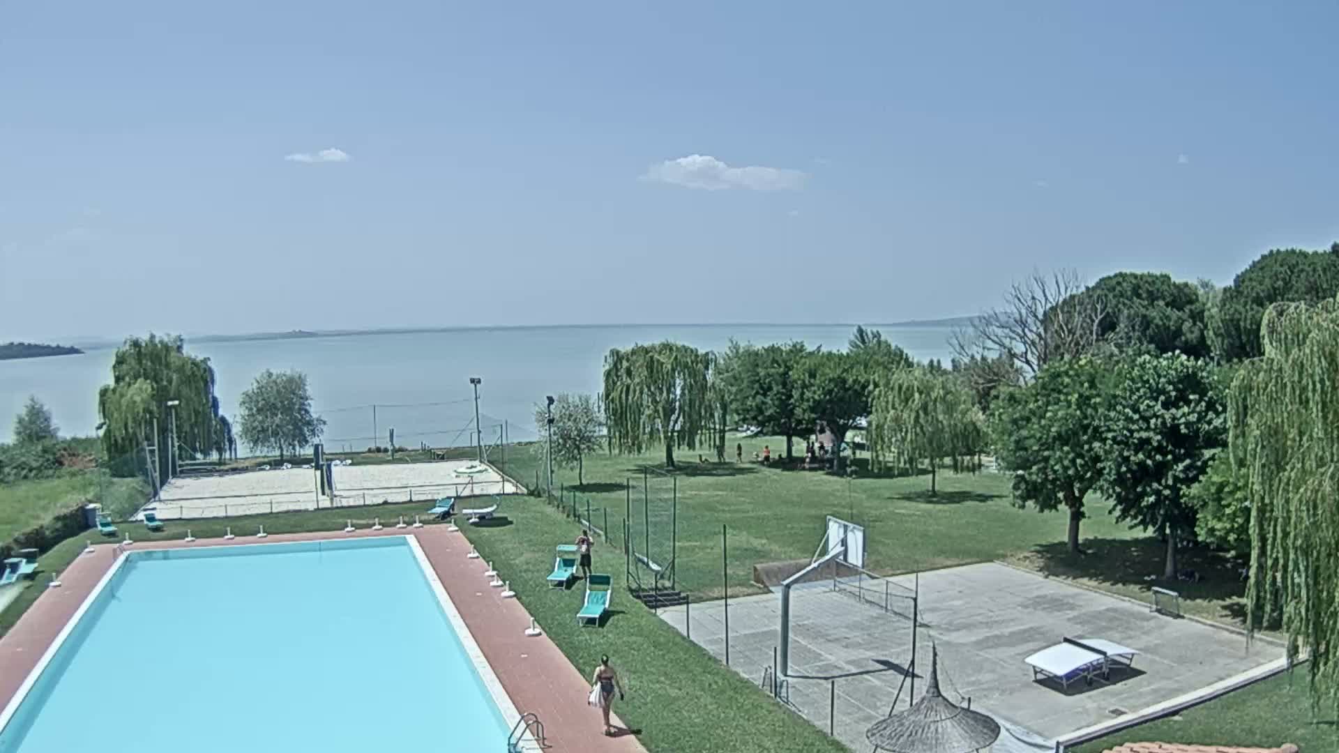A swimming pool, volleyball court, basketball court, and ping pong table are situated on a grassy area overlooking a calm lake under a mostly clear sky.