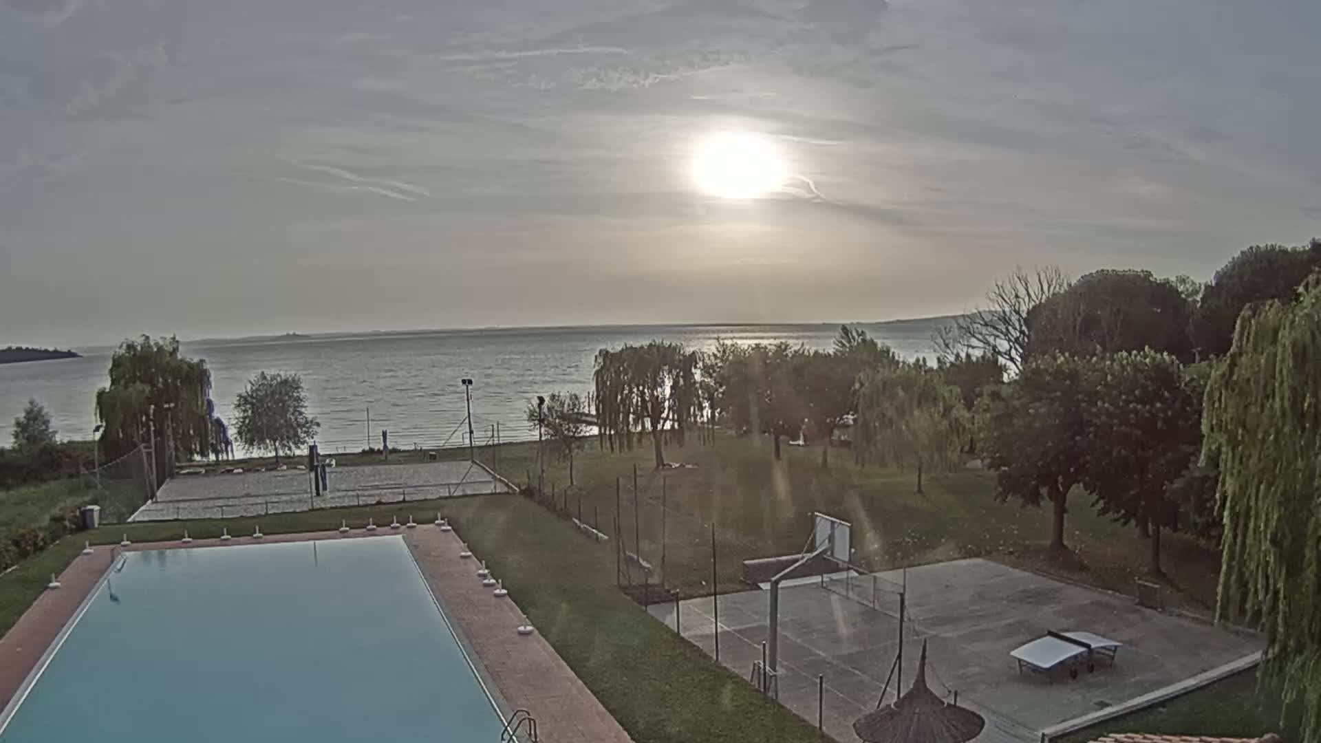 An outdoor scene shows a swimming pool, volleyball court, and basketball court next to a lake under a partly cloudy sky with the sun shining brightly.
