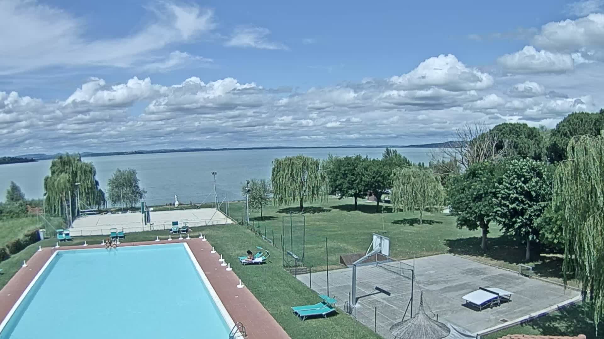 A swimming pool and sports courts are situated on a grassy area overlooking a calm lake under a partly cloudy sky.