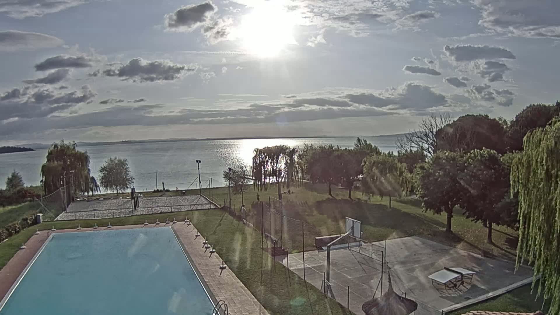 A swimming pool and outdoor recreational areas, including a volleyball court and basketball court, are situated on a grassy expanse overlooking a calm lake under a partly cloudy sky.