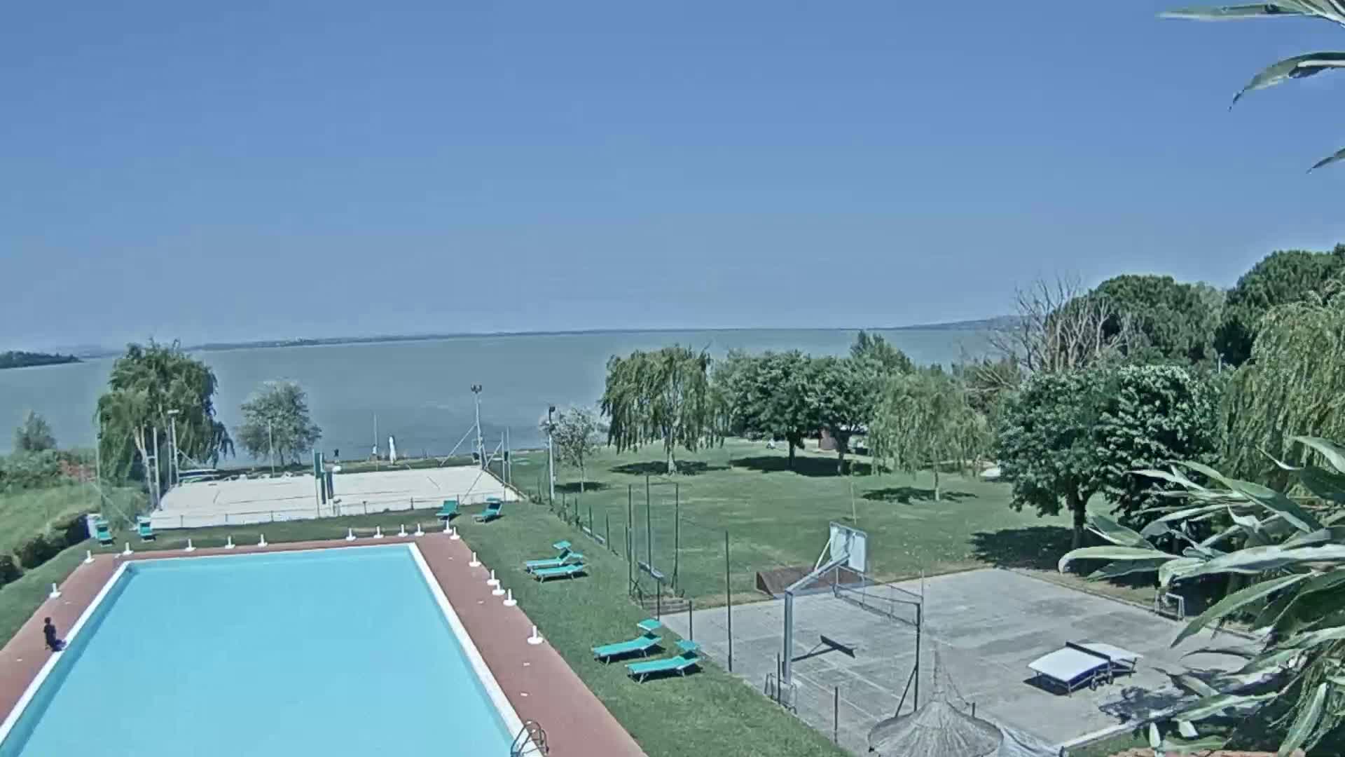 A swimming pool and sports courts are situated on a grassy area overlooking a calm lake under a clear blue sky.