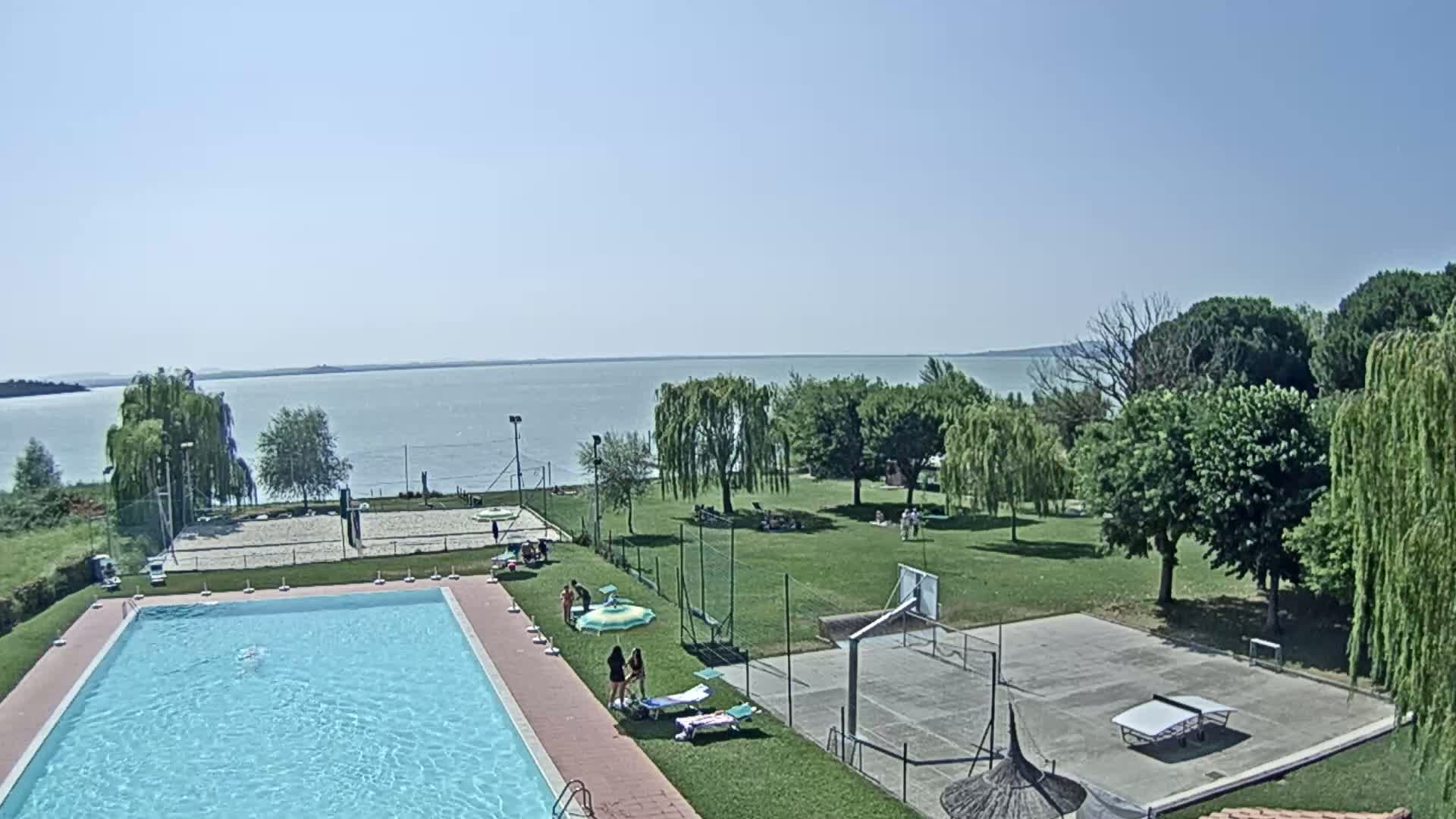 A swimming pool, volleyball court, basketball court, and ping-pong table are situated on a grassy area next to a calm lake on a sunny day.