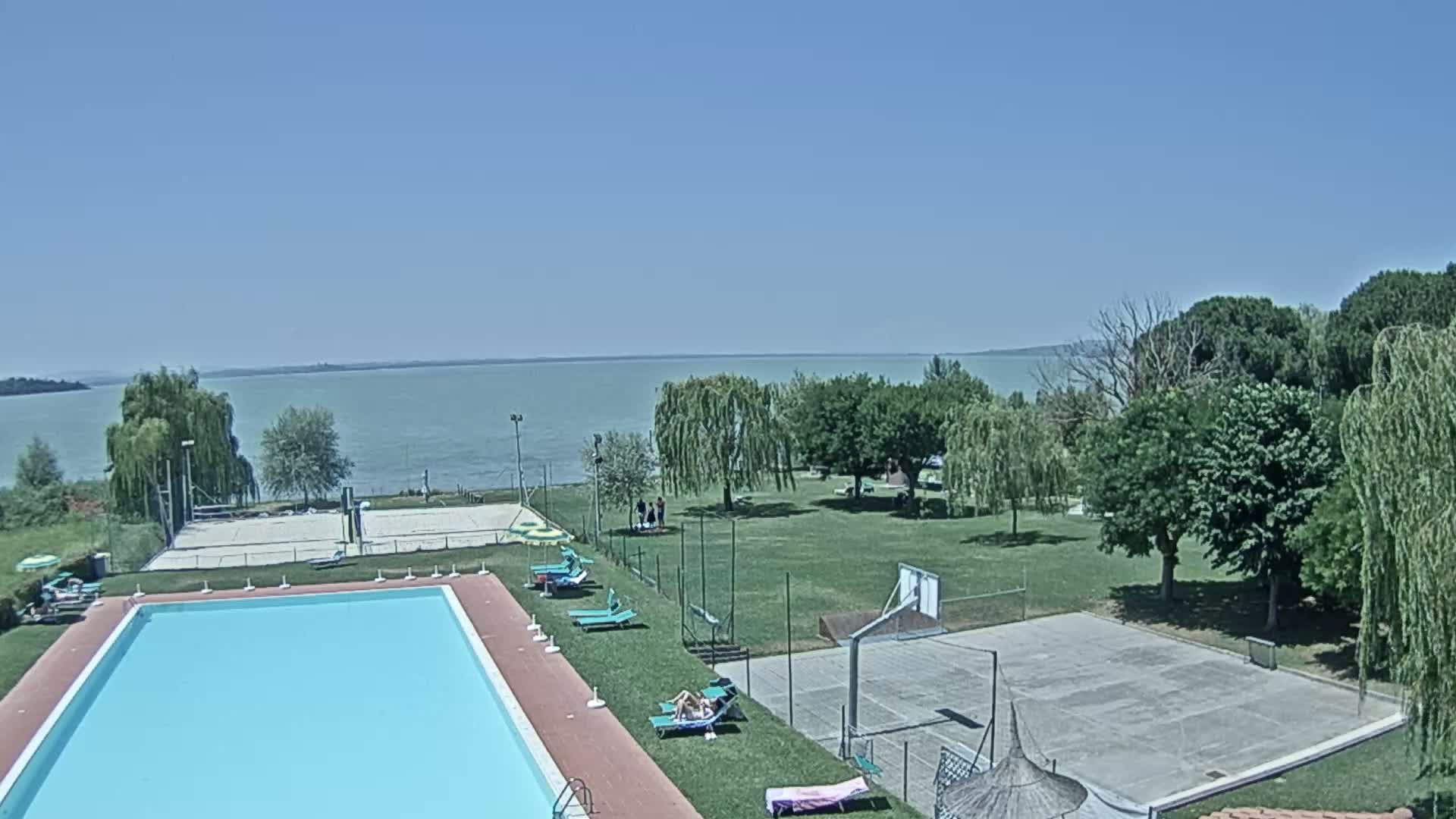 A swimming pool and a basketball court are situated next to a grassy area overlooking a calm lake under a clear blue sky.