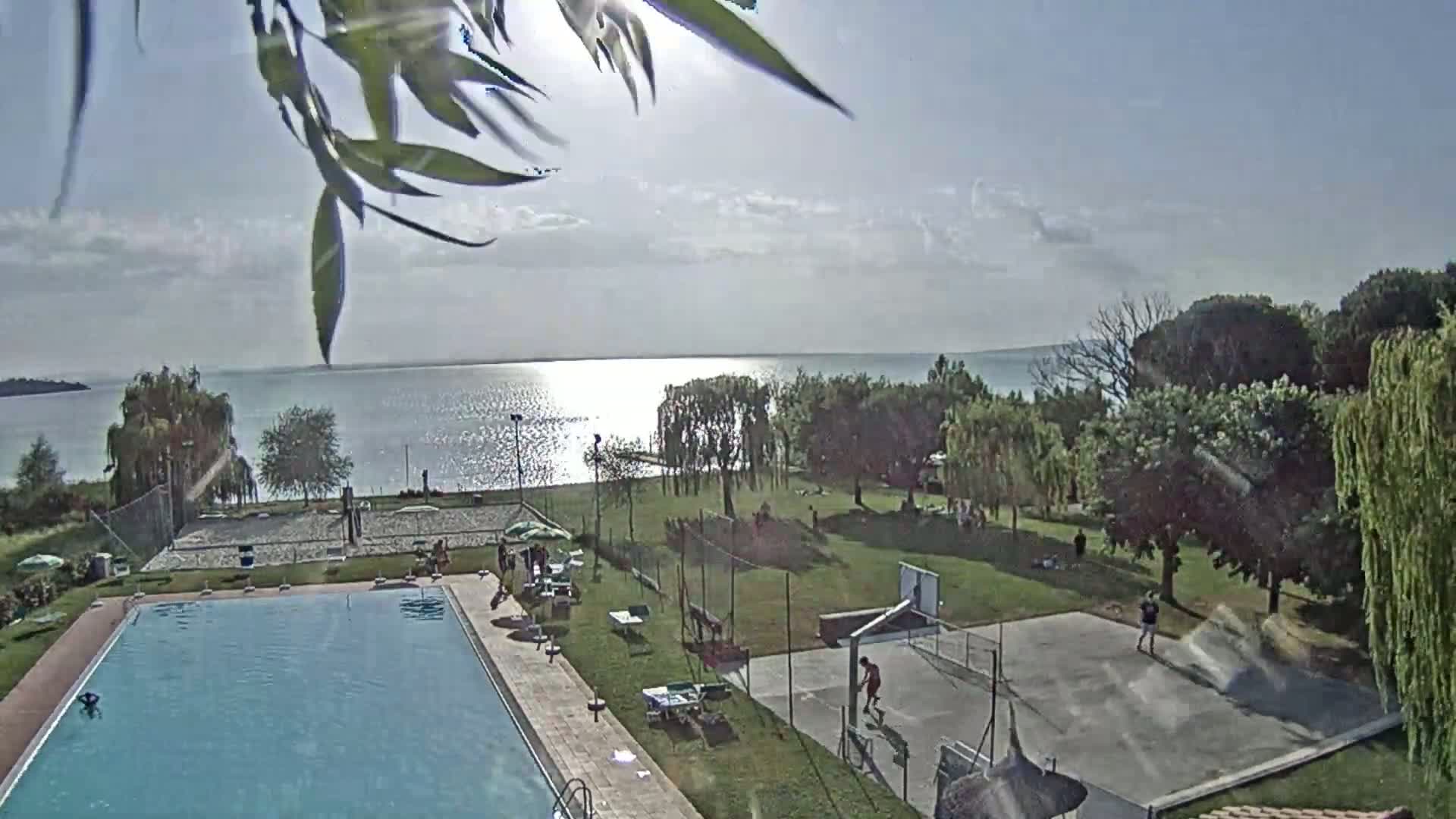 A sunny day overlooks a swimming pool, a volleyball court, a basketball court, and a grassy area next to a calm lake.