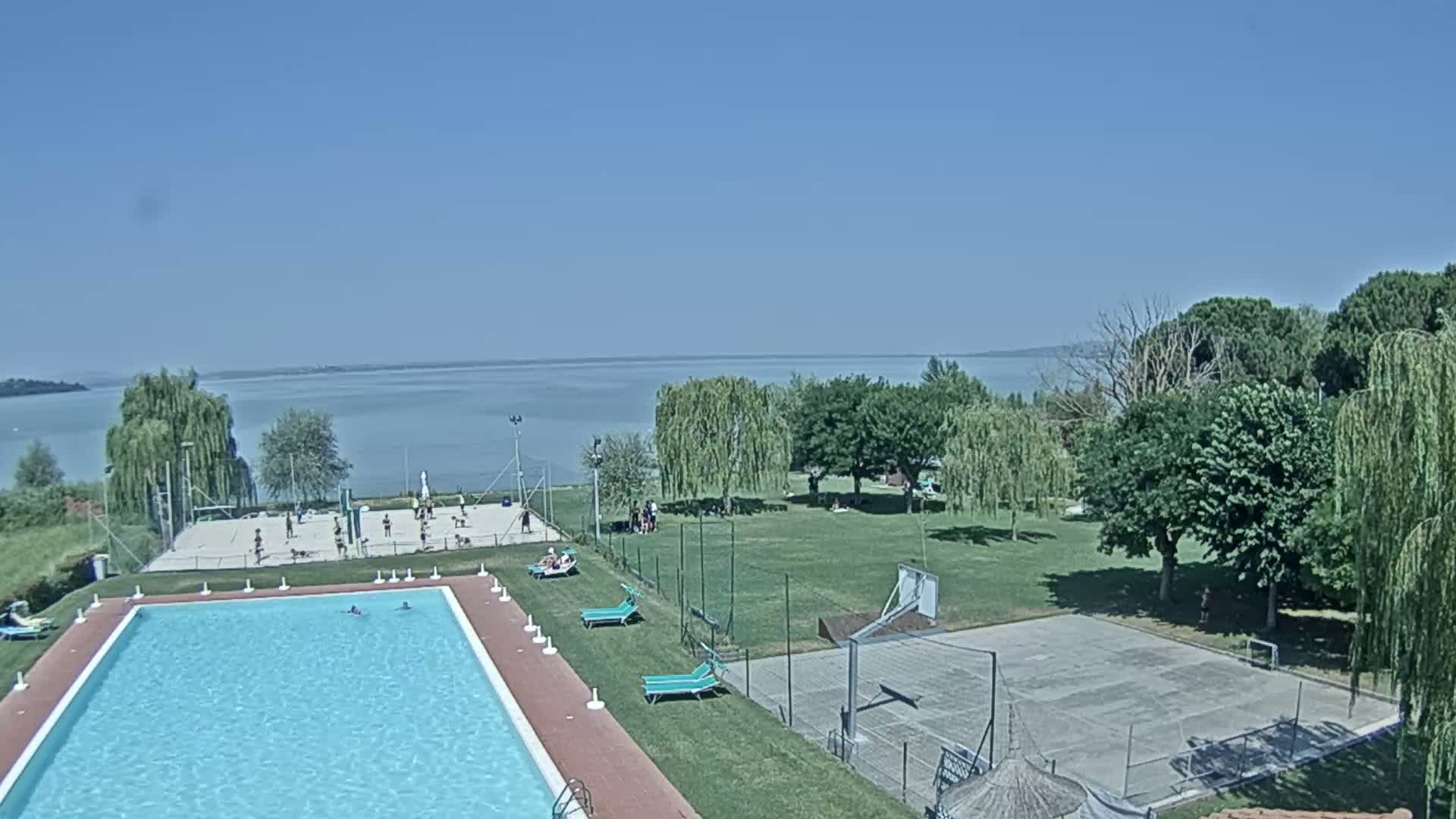 An outdoor swimming pool and recreational area with volleyball and basketball courts are situated near a calm lake under a clear blue sky.