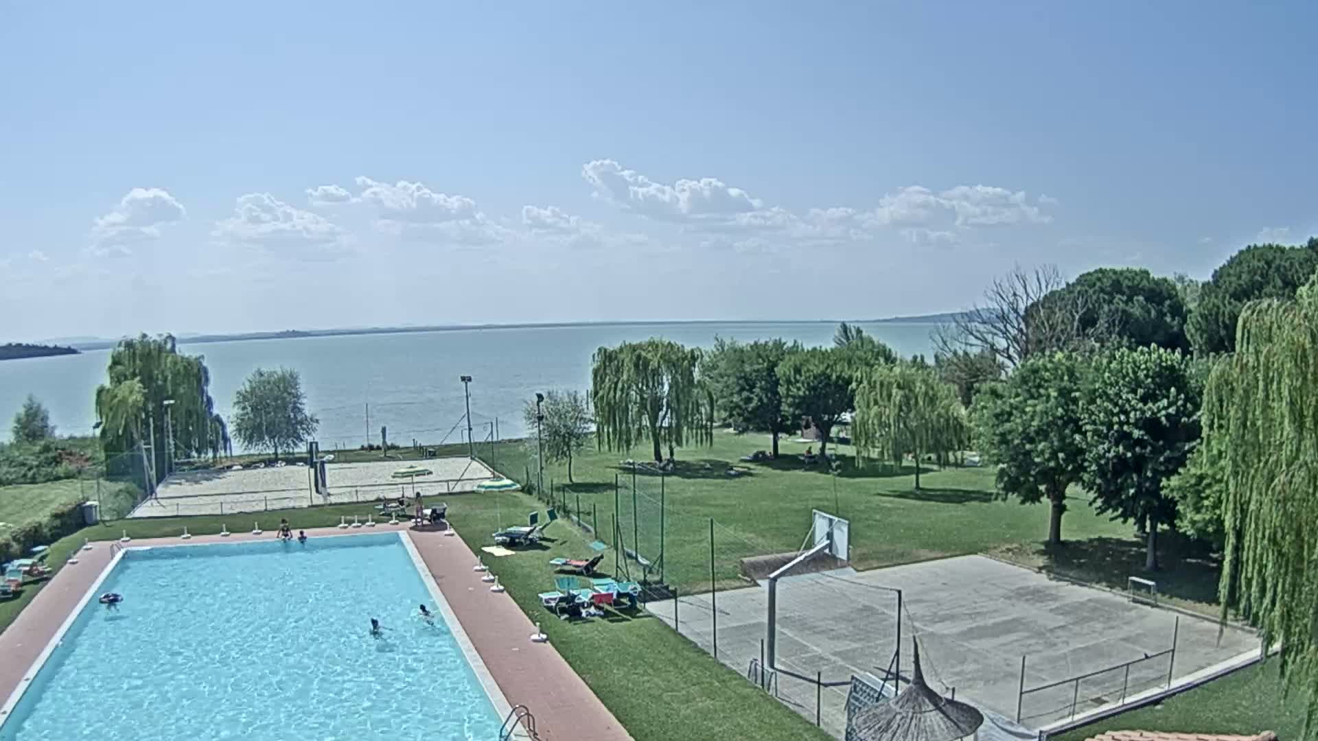 A swimming pool with people swimming is situated beside a basketball court and volleyball court, all overlooking a calm lake under a partly cloudy sky.