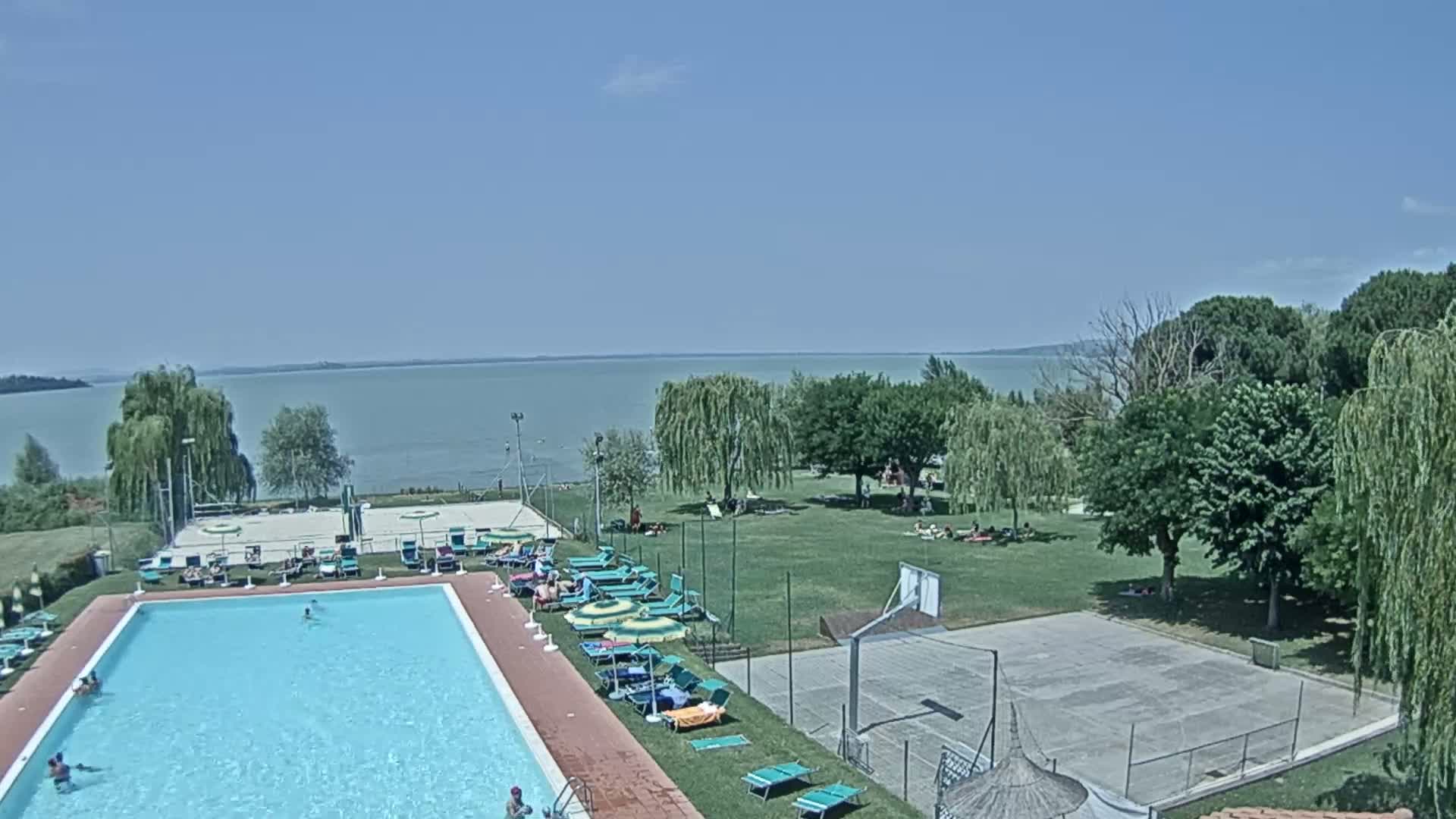 A swimming pool with people swimming and sunbathing is next to a basketball court and grassy area overlooking a calm lake on a clear, sunny day.