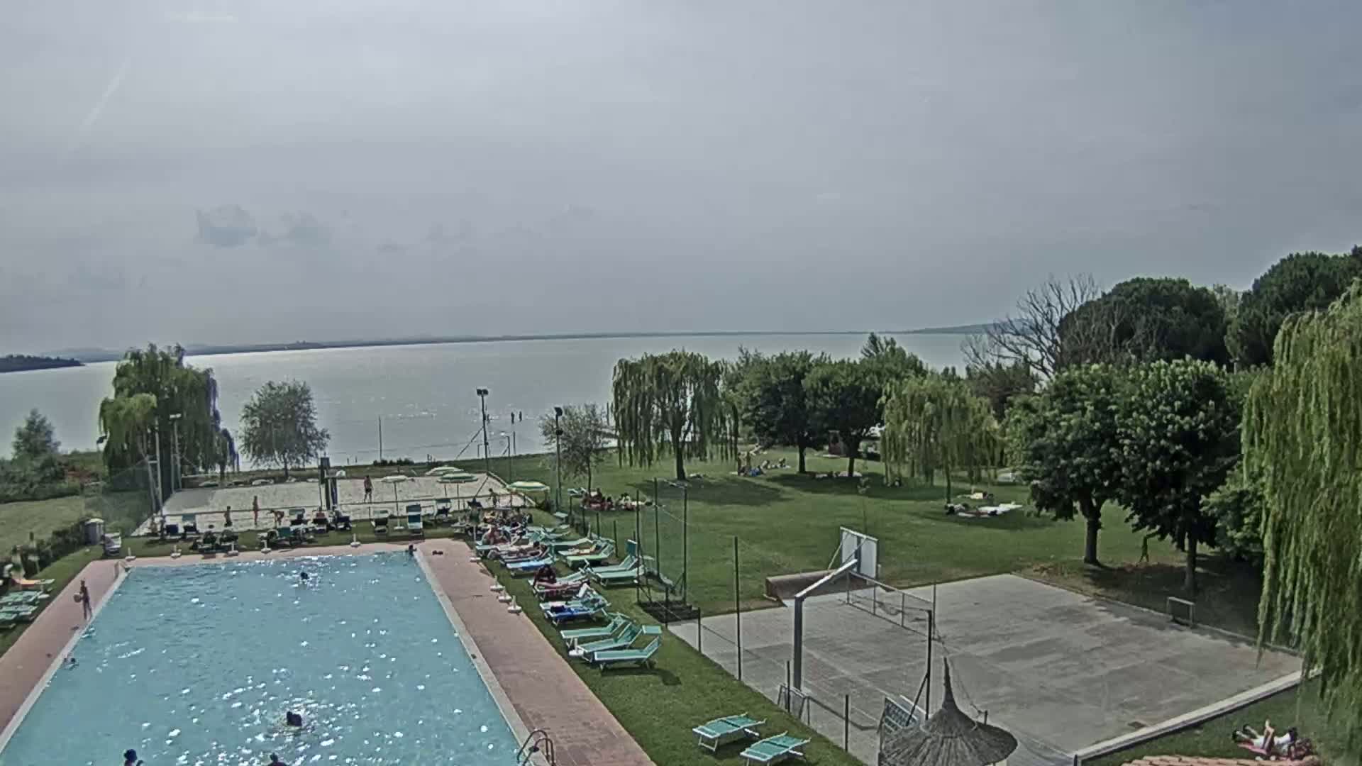 A swimming pool and a basketball court are situated on a grassy area next to a lake, under a partly cloudy sky.
