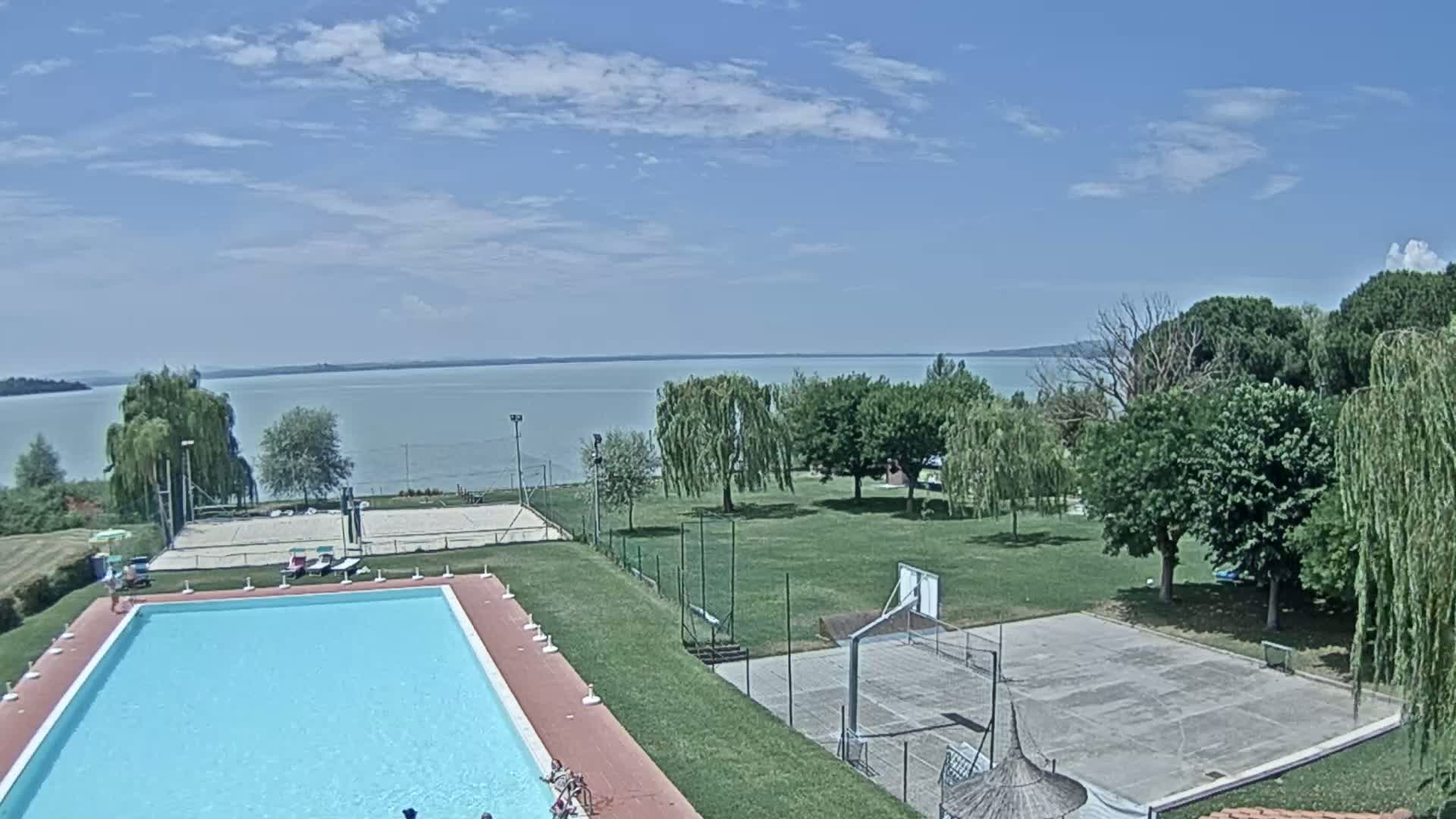A swimming pool and outdoor courts are situated on a grassy area overlooking a calm lake under a partly sunny sky.