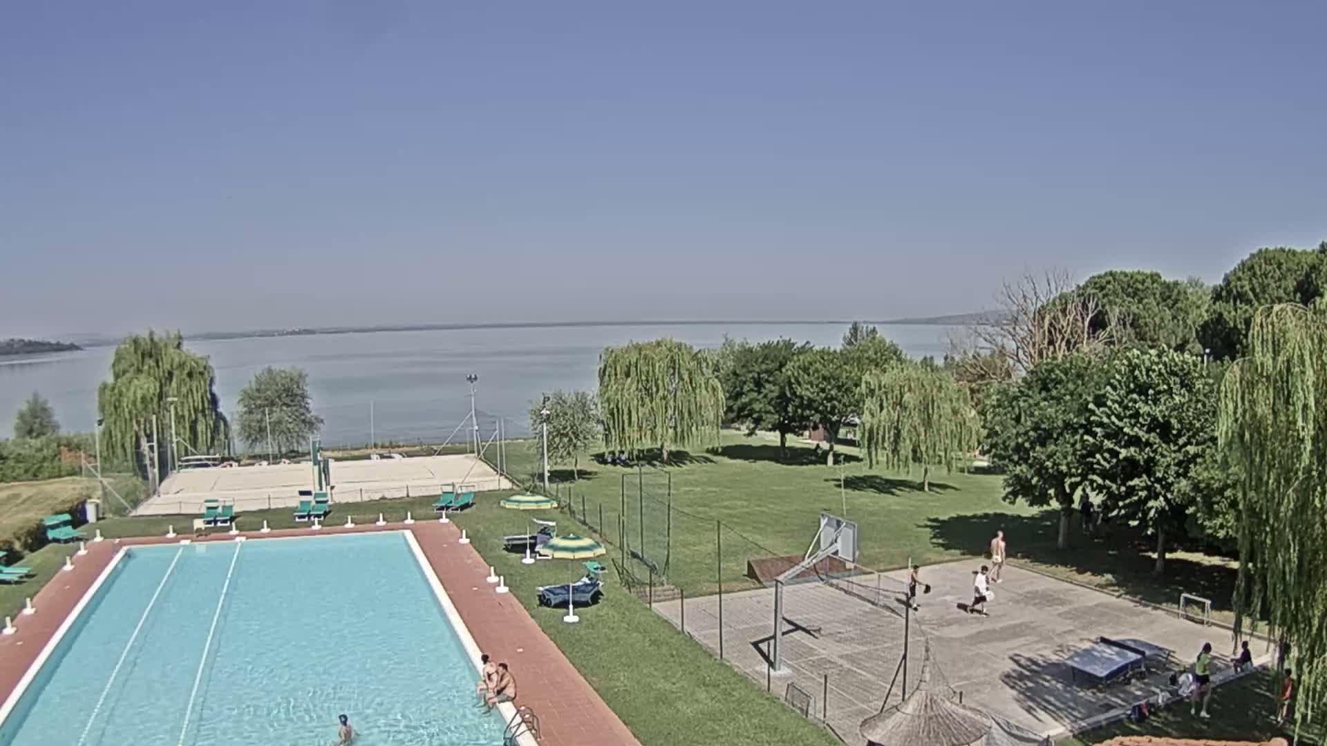 An outdoor swimming pool and sports courts are situated on a grassy area next to a calm lake on a sunny day.