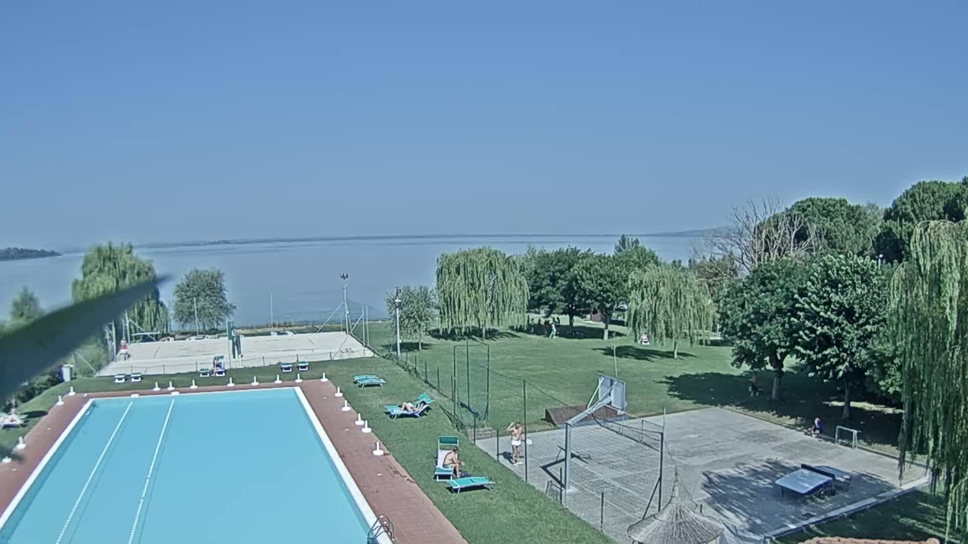 A swimming pool and recreational area with a basketball court and volleyball court are situated near a calm lake on a sunny day.