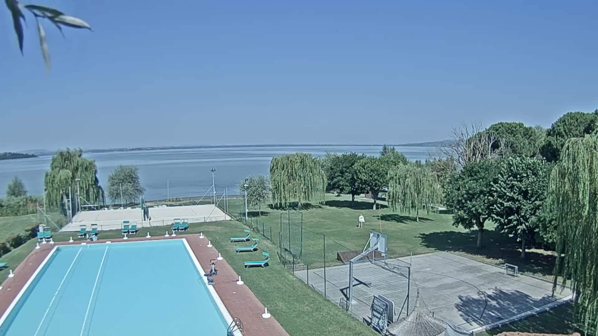An outdoor swimming pool, volleyball court, and basketball court are situated near a calm lake on a sunny day.