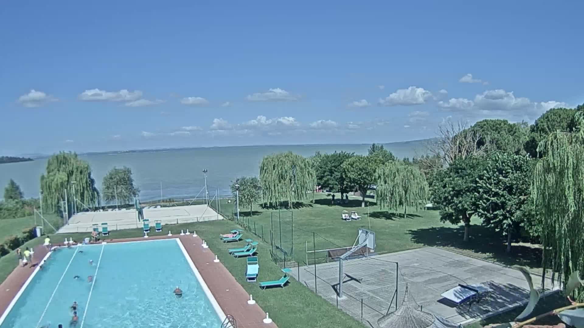 A swimming pool with people swimming in it is next to a basketball court and volleyball court, all situated on a grassy area overlooking a calm lake under a mostly sunny sky.