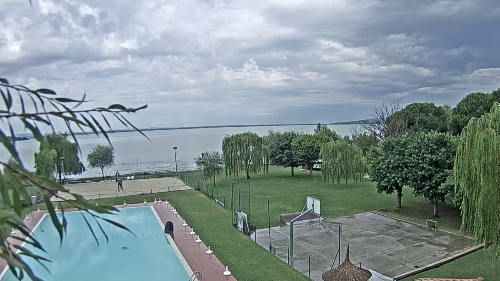 A swimming pool and outdoor sports courts are situated on a grassy area overlooking a calm lake under a cloudy sky.