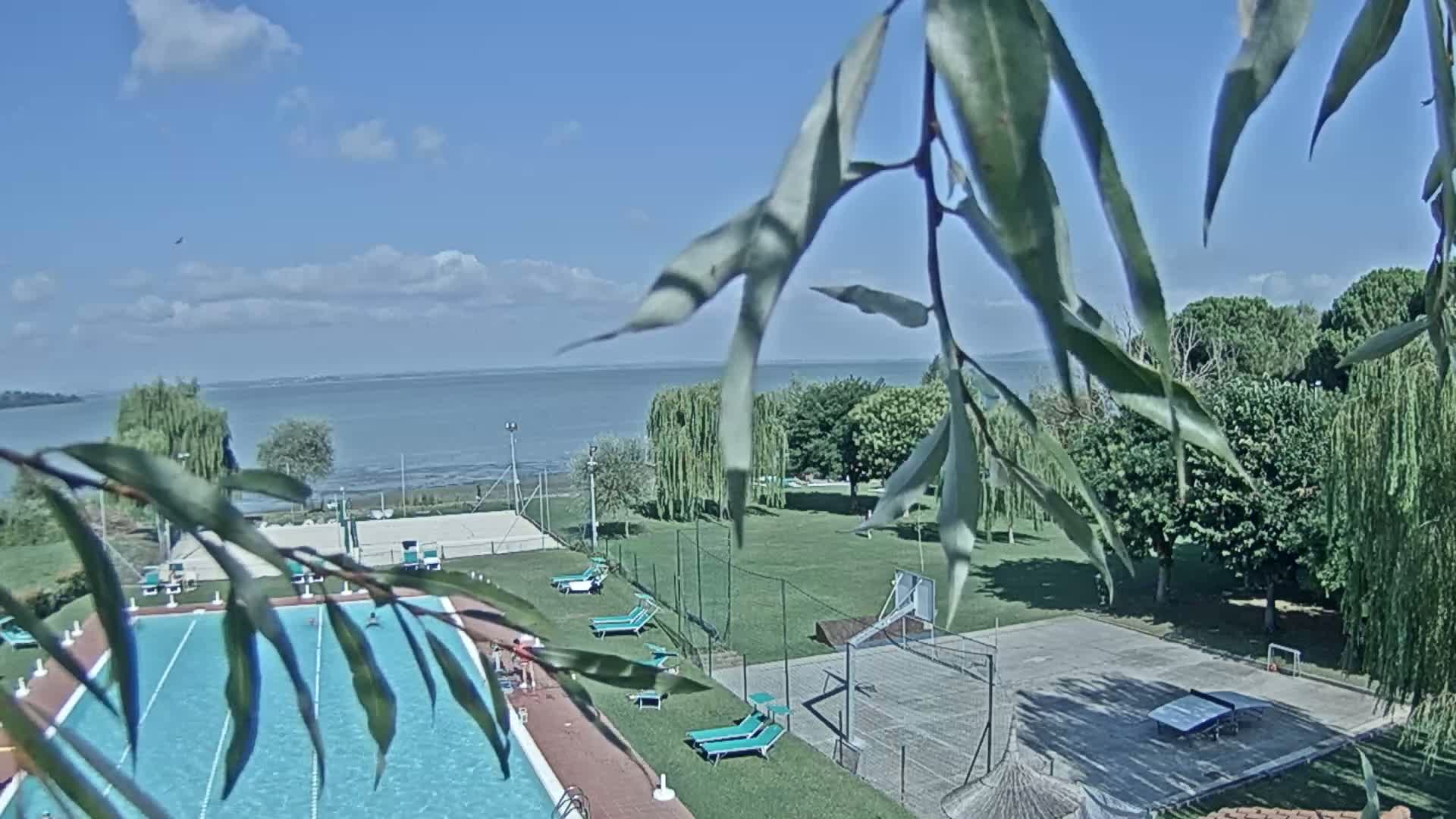 An outdoor swimming pool and recreational area with tennis and basketball courts are situated beside a calm lake under a partly sunny sky.