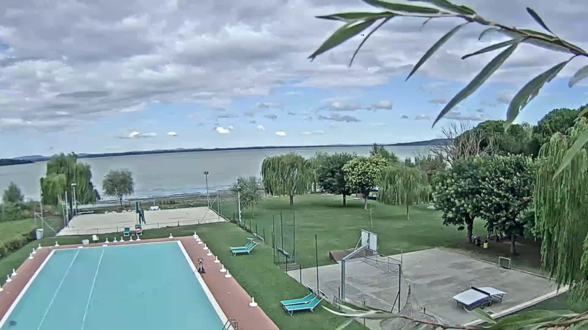 A swimming pool and several outdoor courts are situated on a grassy area overlooking a calm lake under a partly cloudy sky.