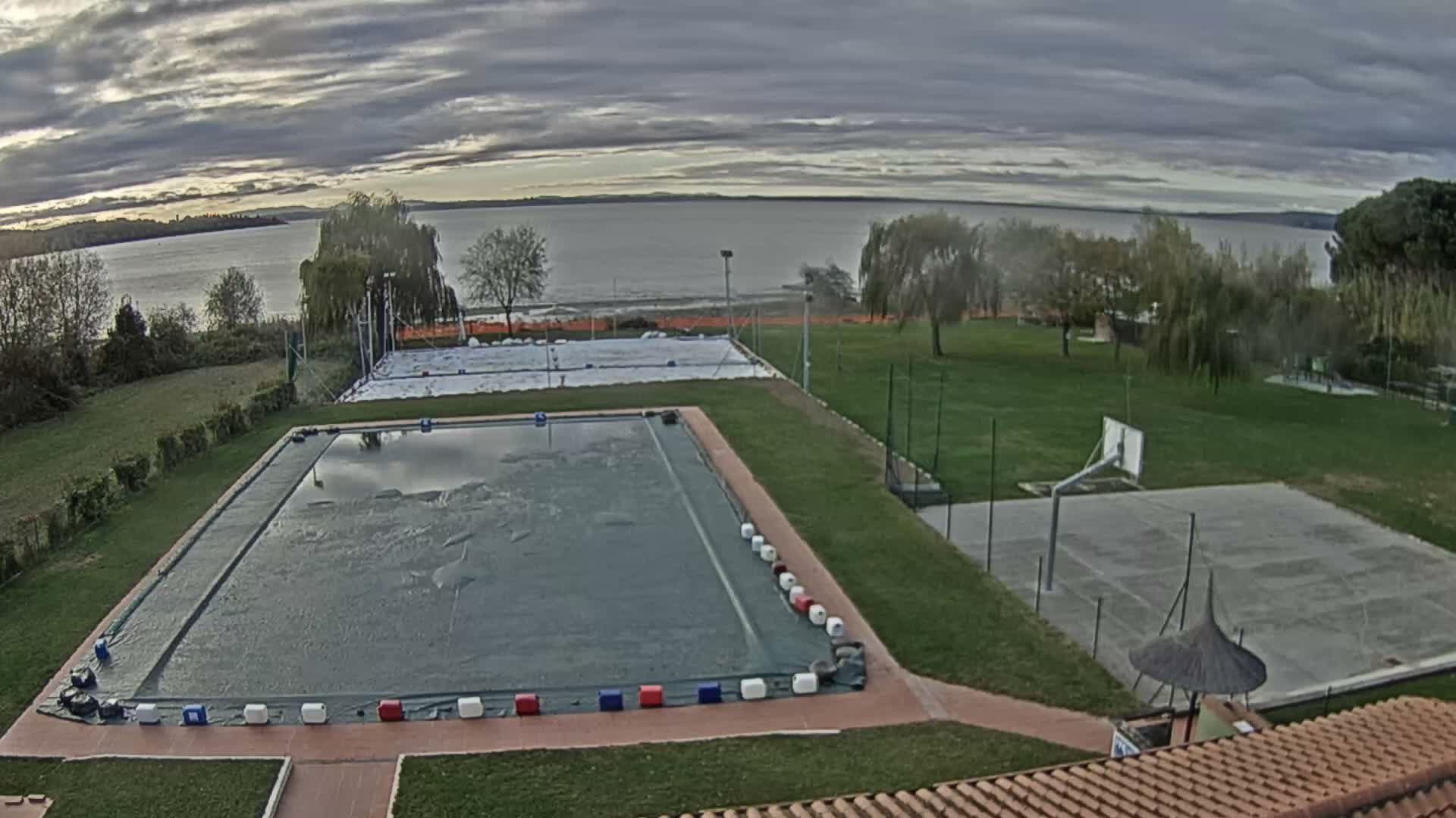 A swimming pool and several outdoor courts are situated on a grassy area overlooking a calm lake under a partly cloudy sky.