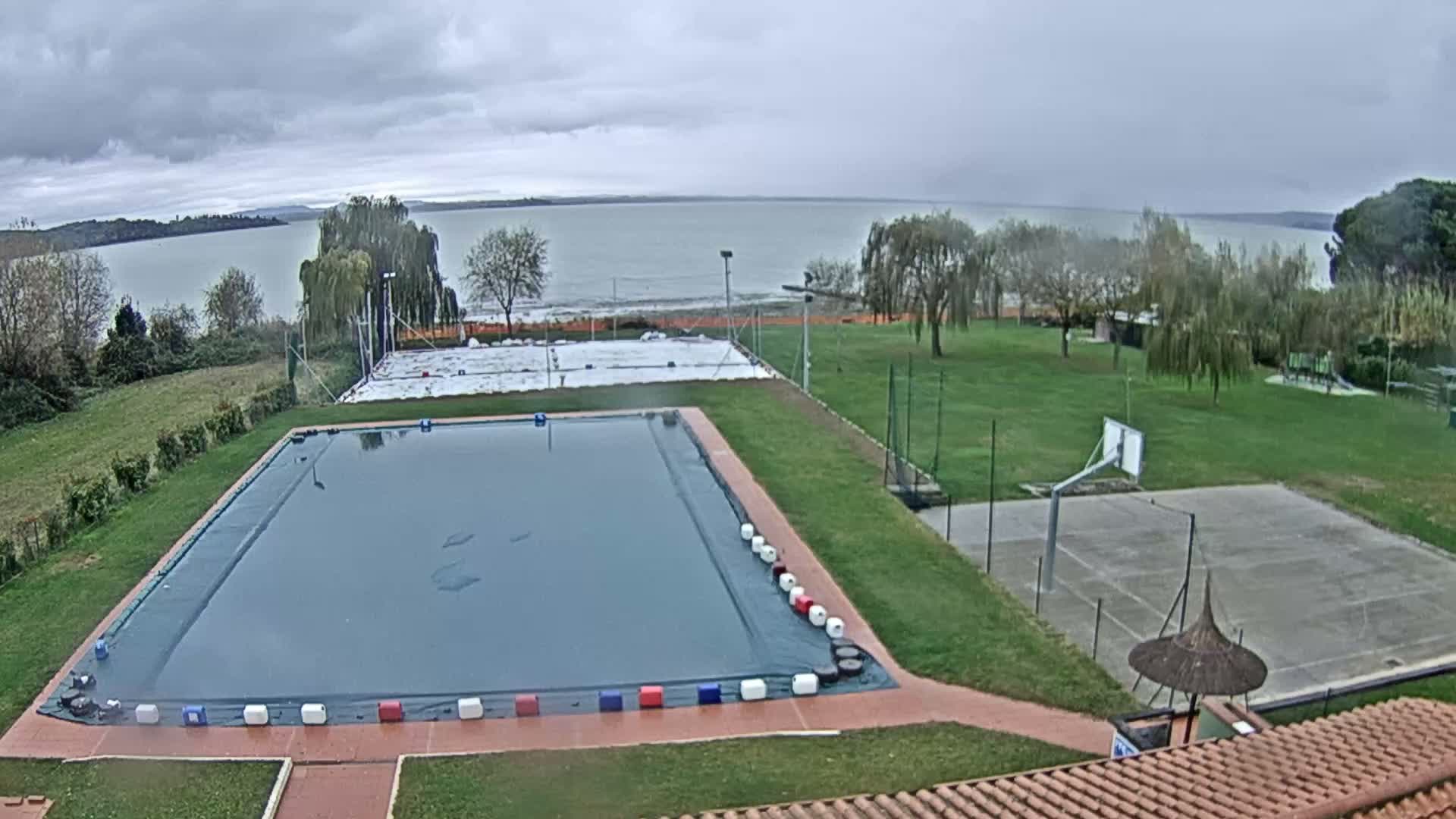 On an overcast day, a large covered swimming pool, a green lawn, and a basketball court are visible in the foreground, with a white-tarped court, more green space, and a lake lined by trees in the background.