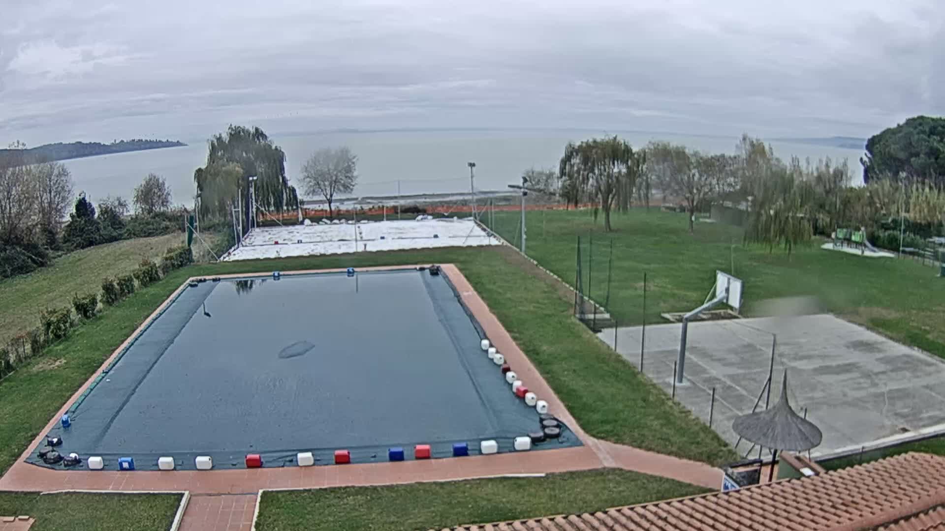 Under an overcast sky, a large covered swimming pool, a basketball court, and a white-covered tennis court are situated on green grounds dotted with trees, overlooking a wide body of water with distant land.