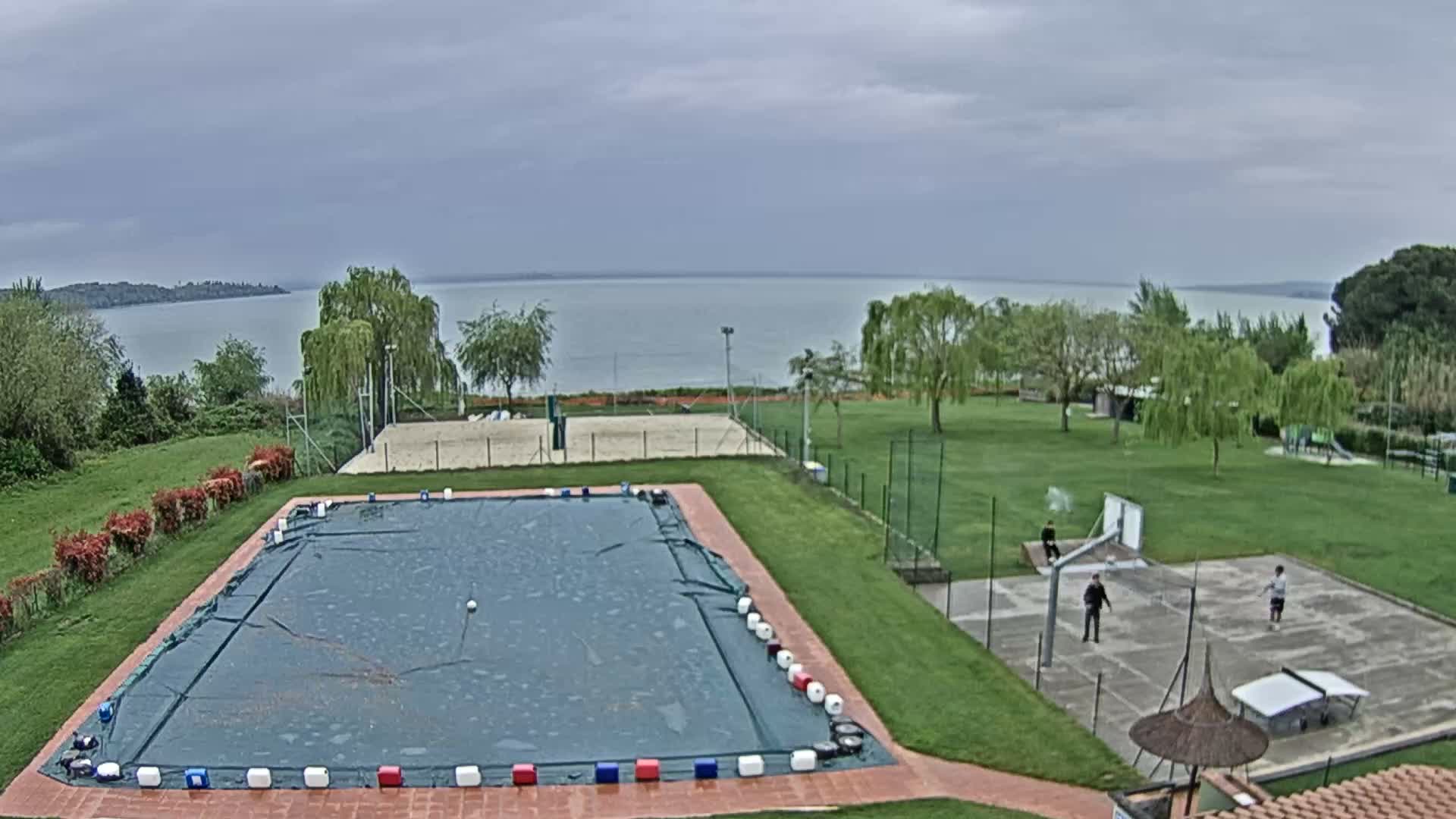 An outdoor scene features a large covered swimming pool, a concrete basketball court, and grassy lawns with scattered trees, all visible under a dull, overcast sky with a misty distant landscape and patches of snow on a far field.