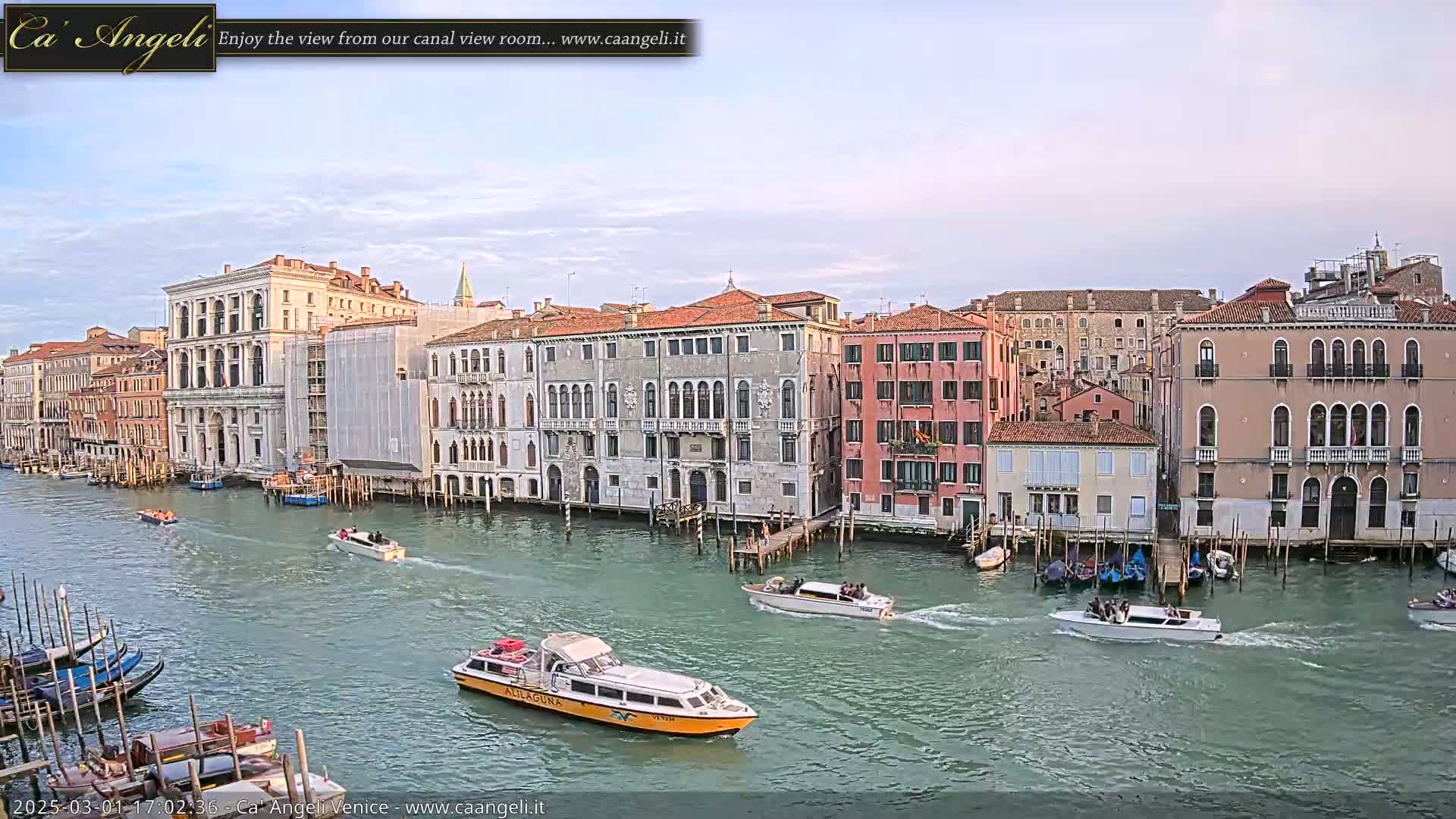 Several boats travel along a canal in Venice, lined with tall, historic buildings under a partly cloudy sky.