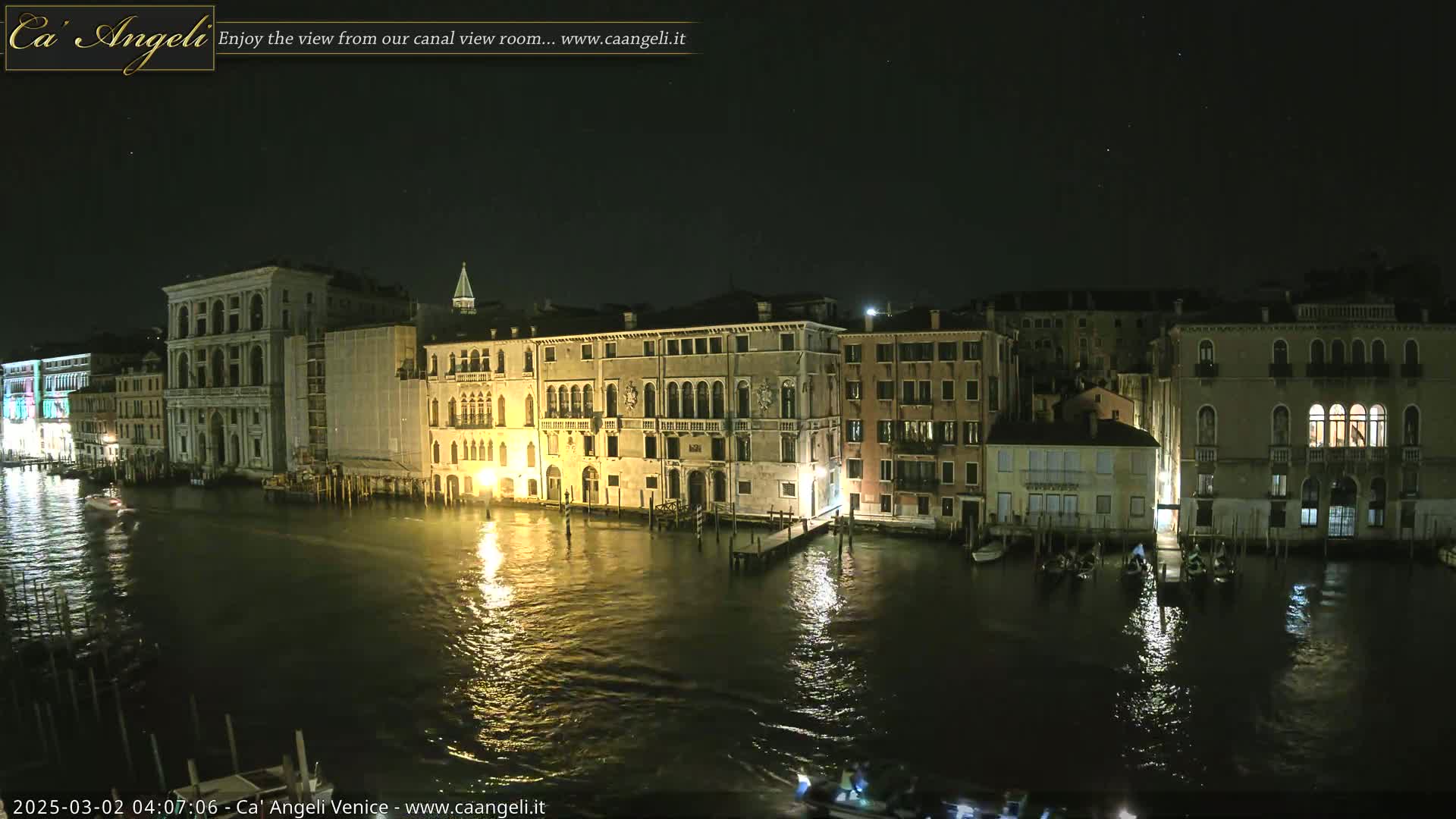 A nighttime view of a canal in Venice, Italy, shows several illuminated buildings lining the waterway, with boats moored along the edges under a clear, starry sky.
