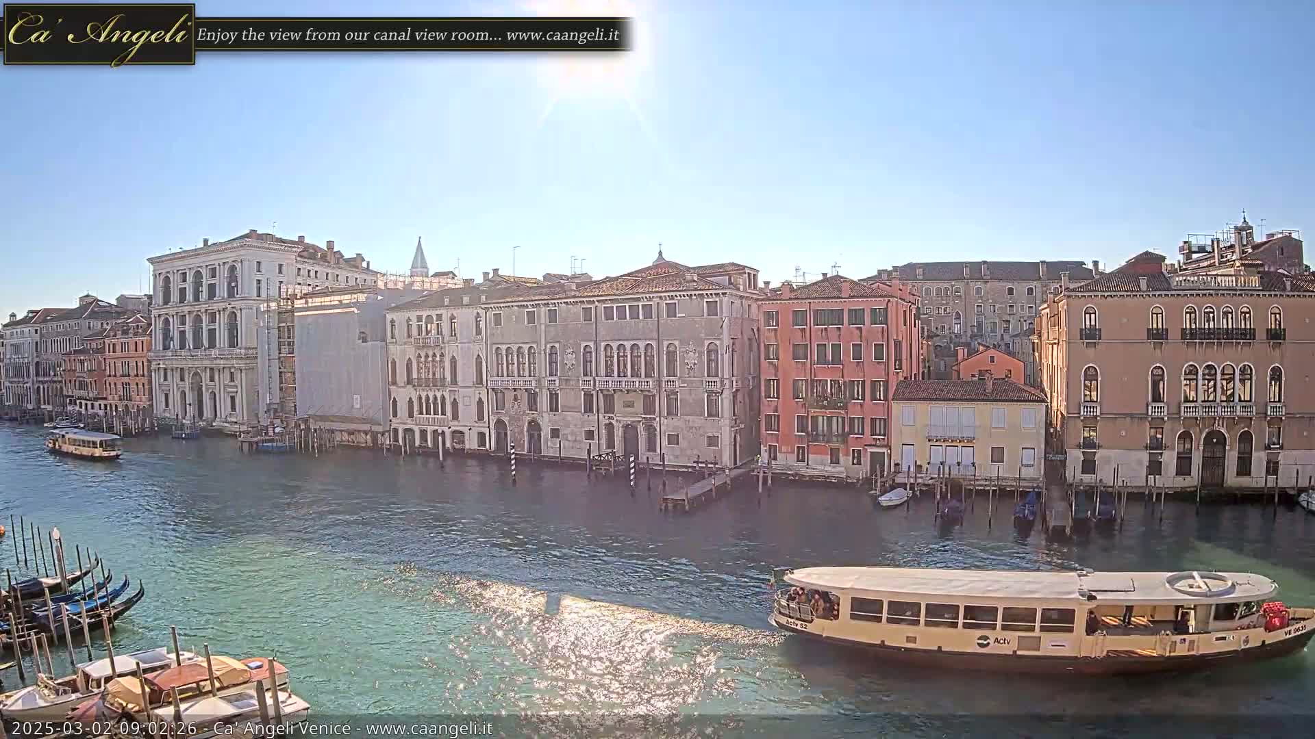 A sunny day in Venice shows a canal with several boats, gondolas, and buildings lining the waterway.