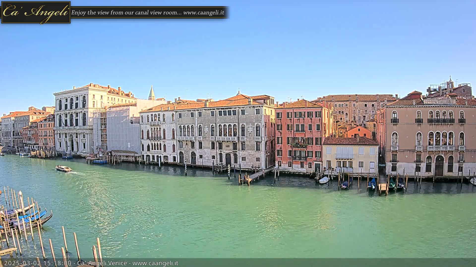 A canal in Venice, under a clear blue sky, is lined with several historic buildings, with gondolas moored along the waterway and a motorboat passing by.
