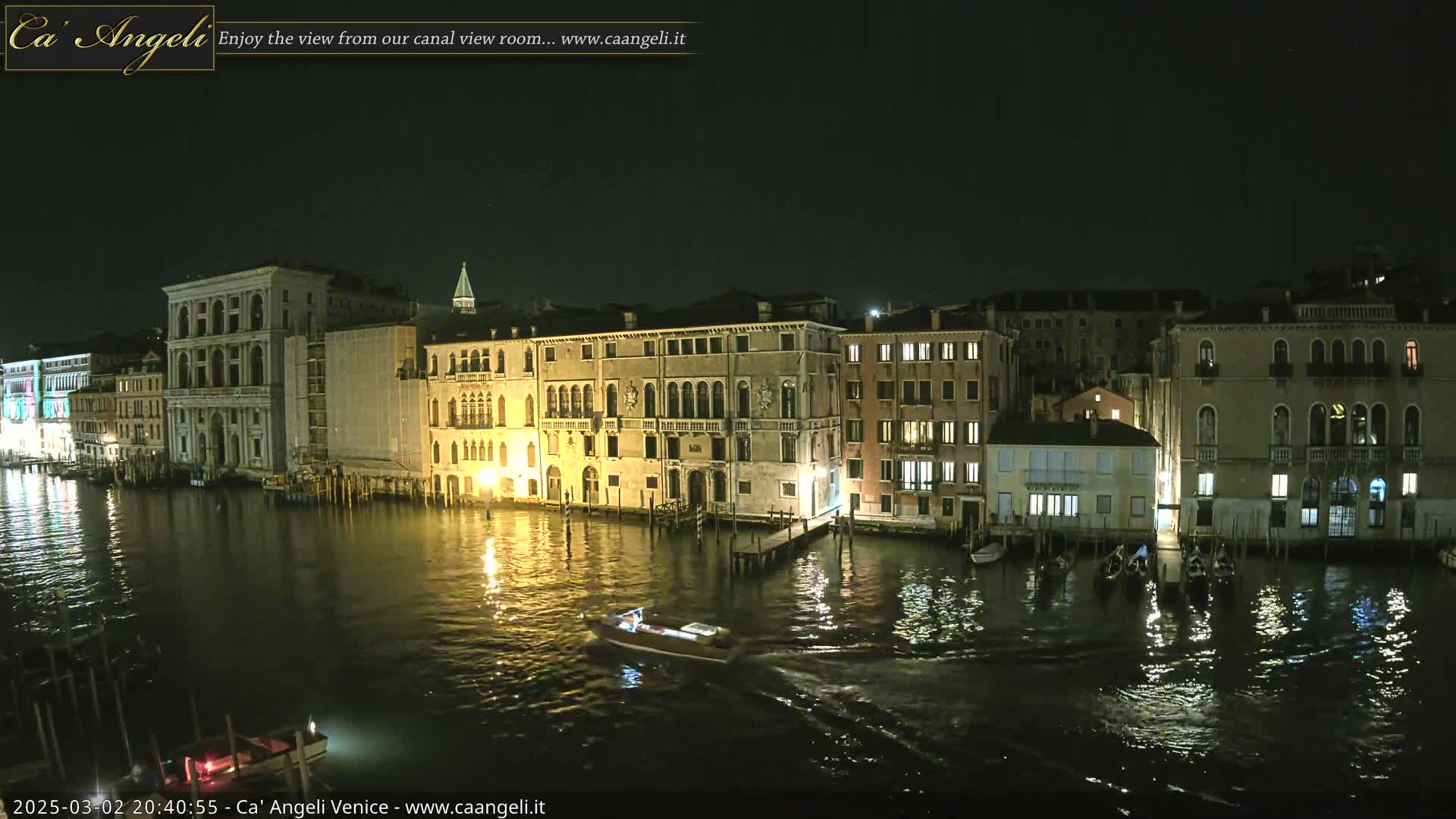 At night, a canal in Venice shows several buildings lit along its banks with a motorboat and gondolas on the water.