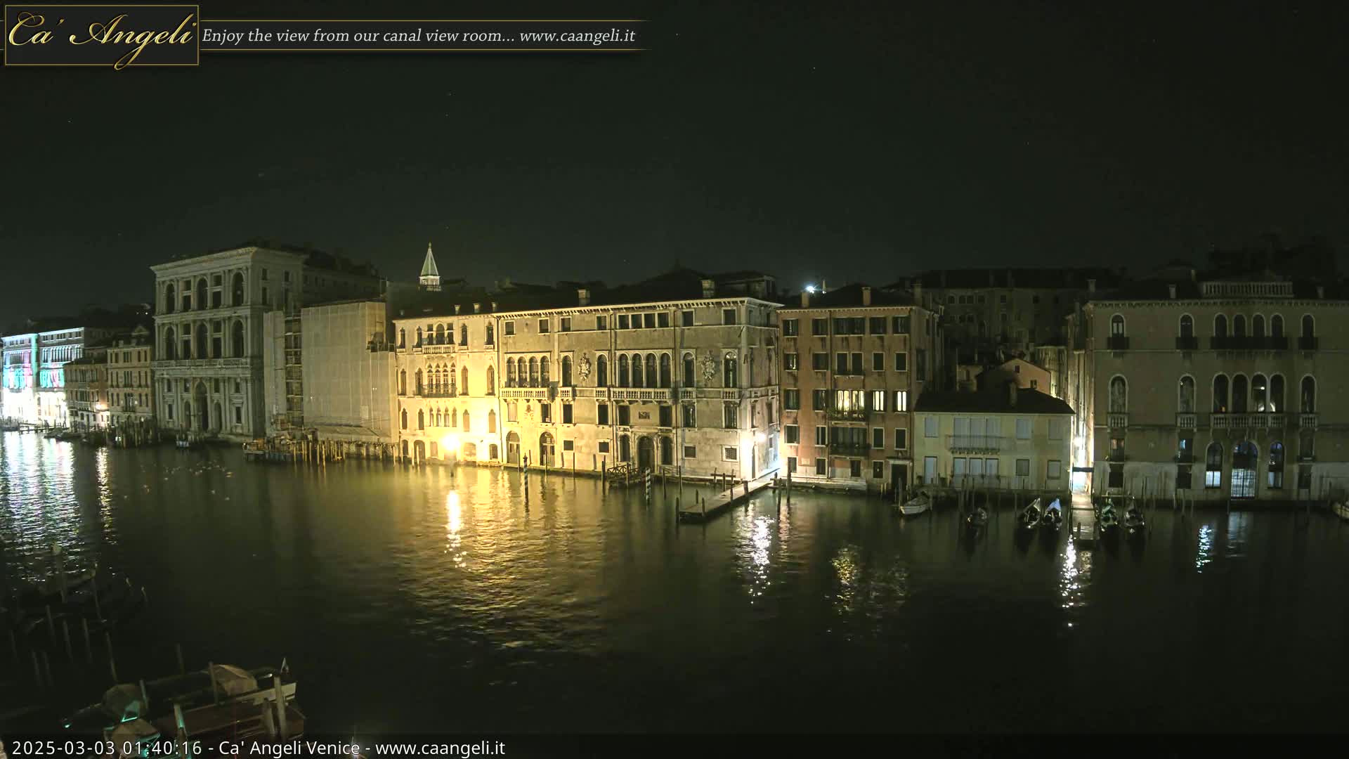 A nighttime view of a canal in Venice, showing several illuminated buildings along the water's edge with gondolas moored nearby under a clear, dark sky.