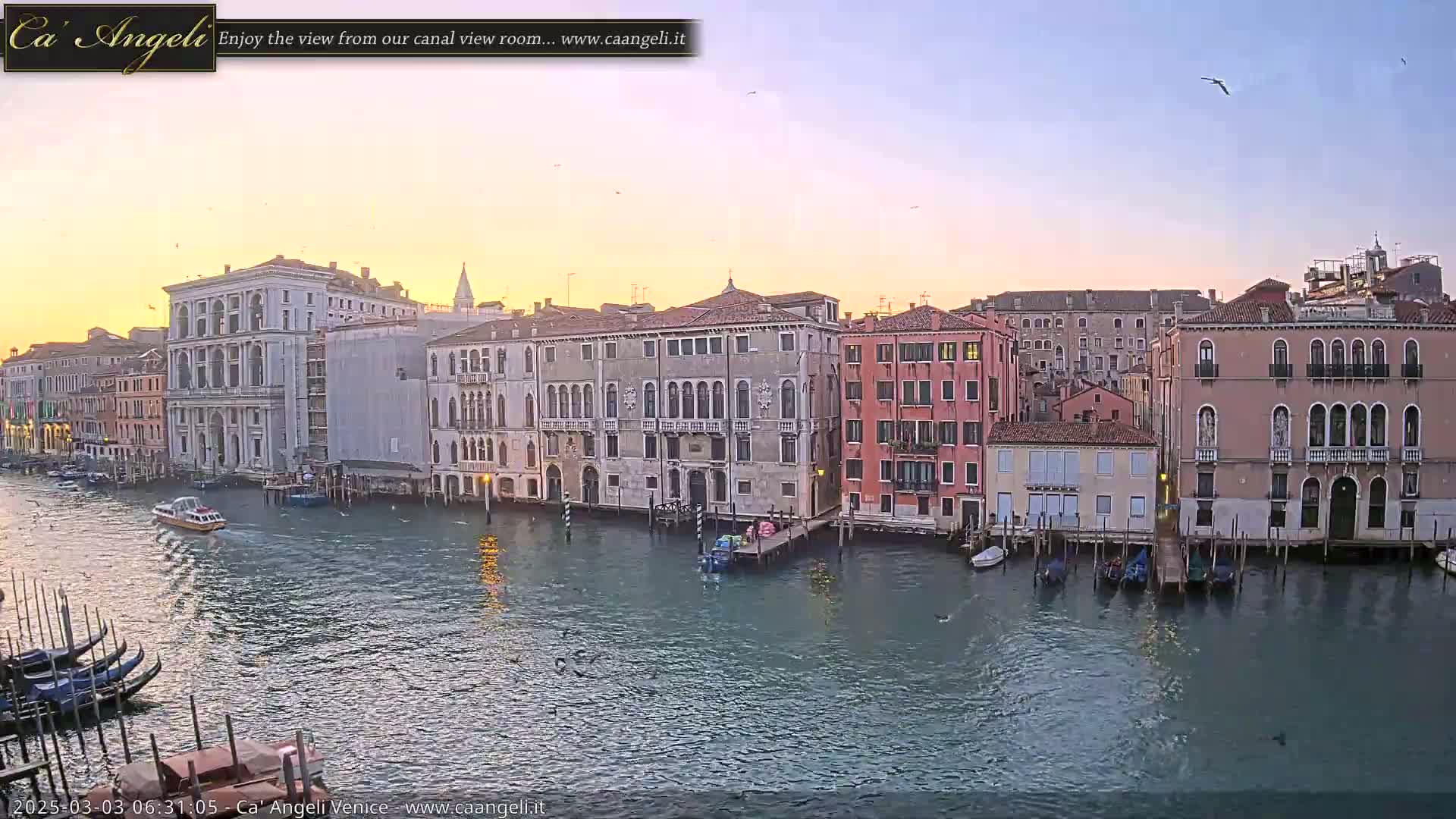 A calm canal in Venice at sunrise, lined with historic buildings, with several boats on the water.