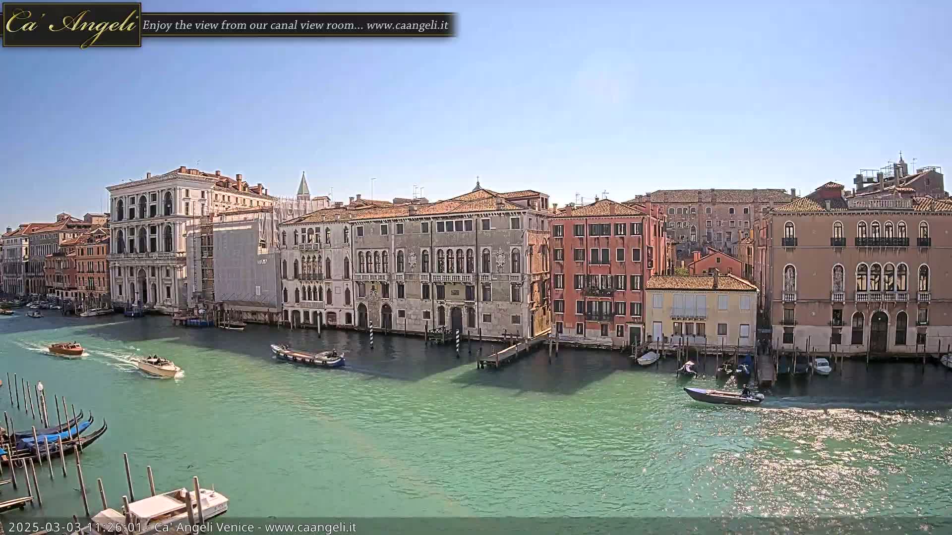 A sunny day in Venice shows several gondolas and motorboats on a canal lined with historic buildings.