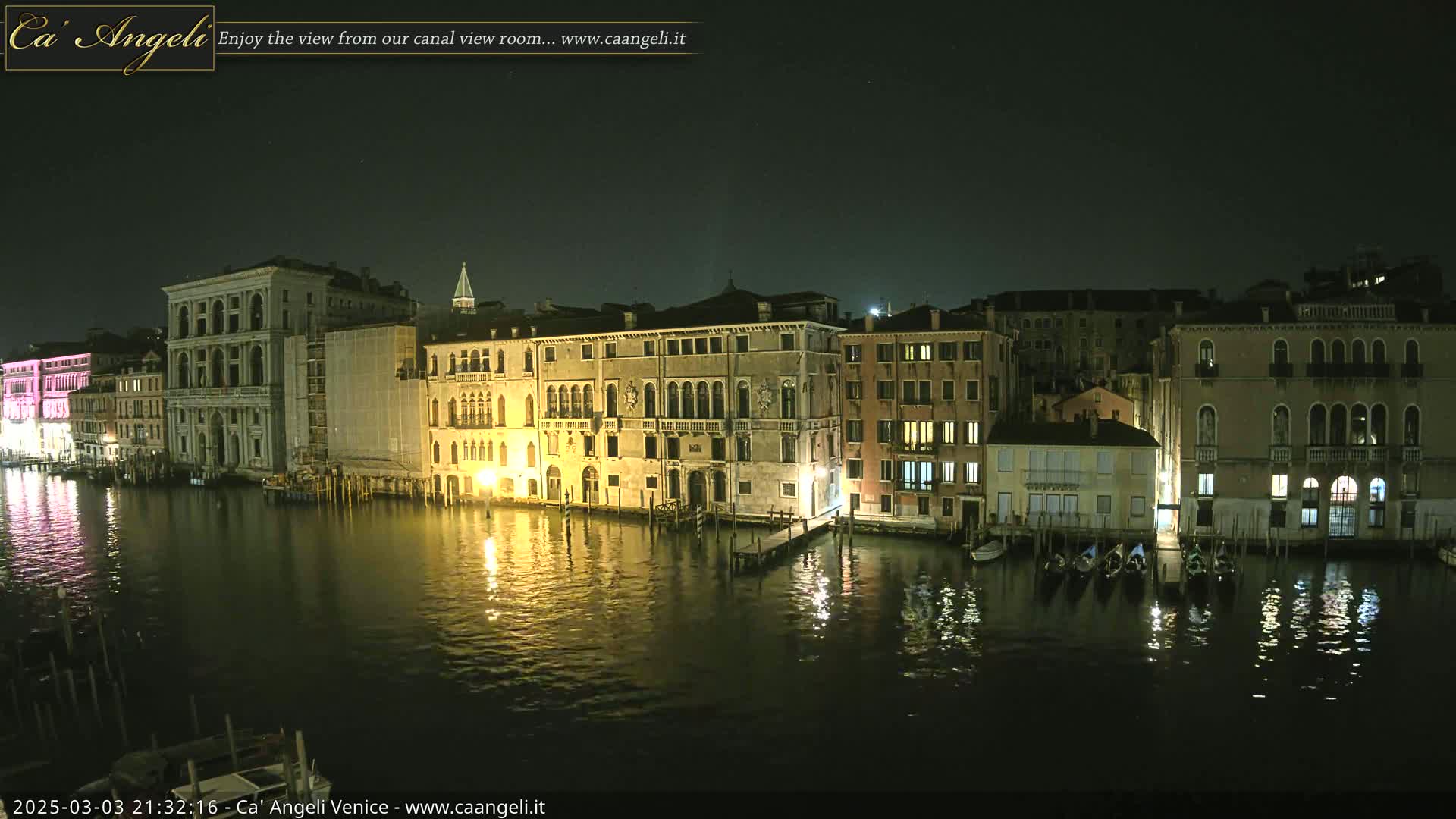 A nighttime view of a canal in Venice, Italy, shows several illuminated buildings lining the waterway, with gondolas moored along the banks.