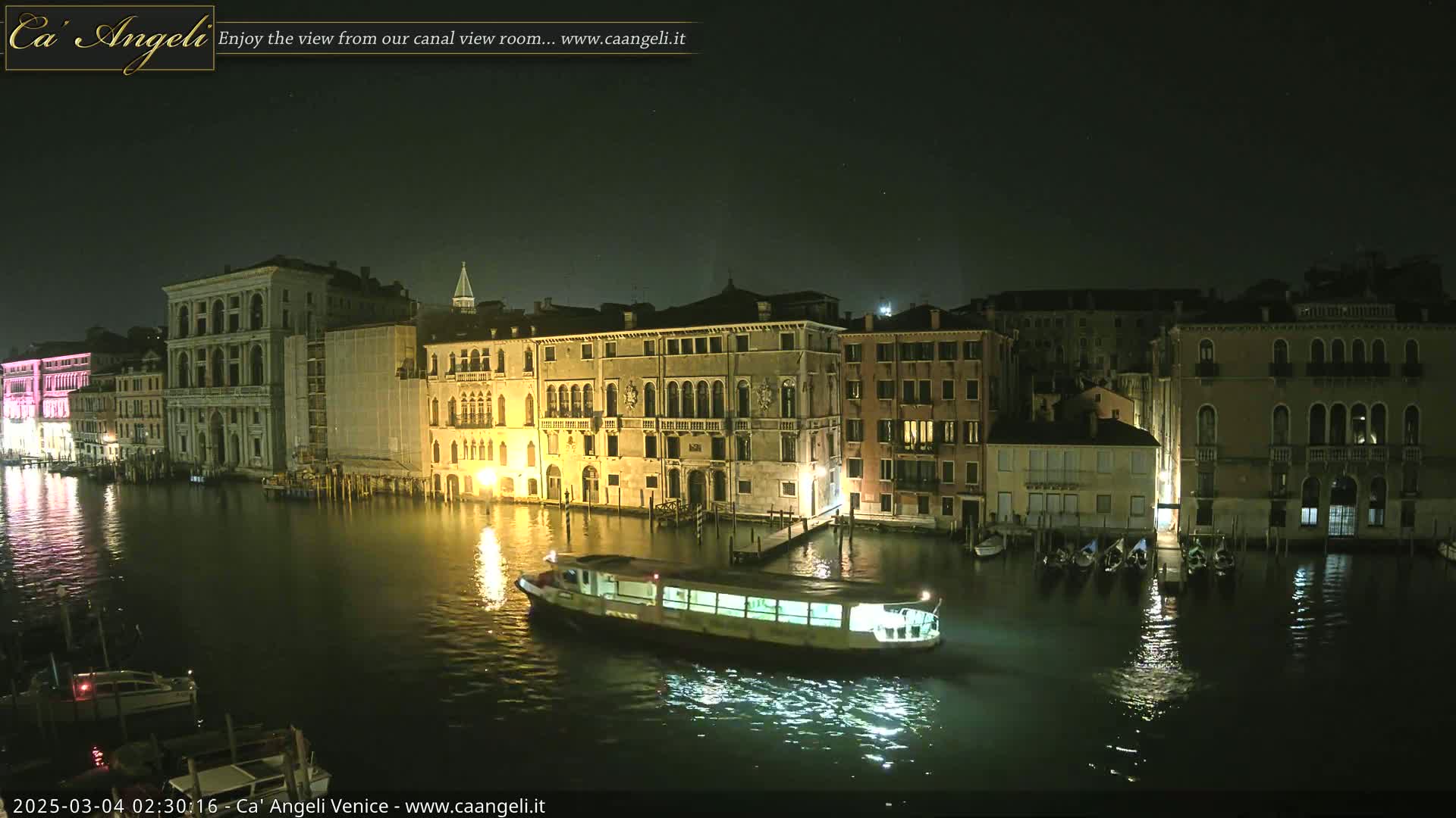 A nighttime view of a canal in Venice, with several illuminated buildings lining the waterway and a boat on the water.