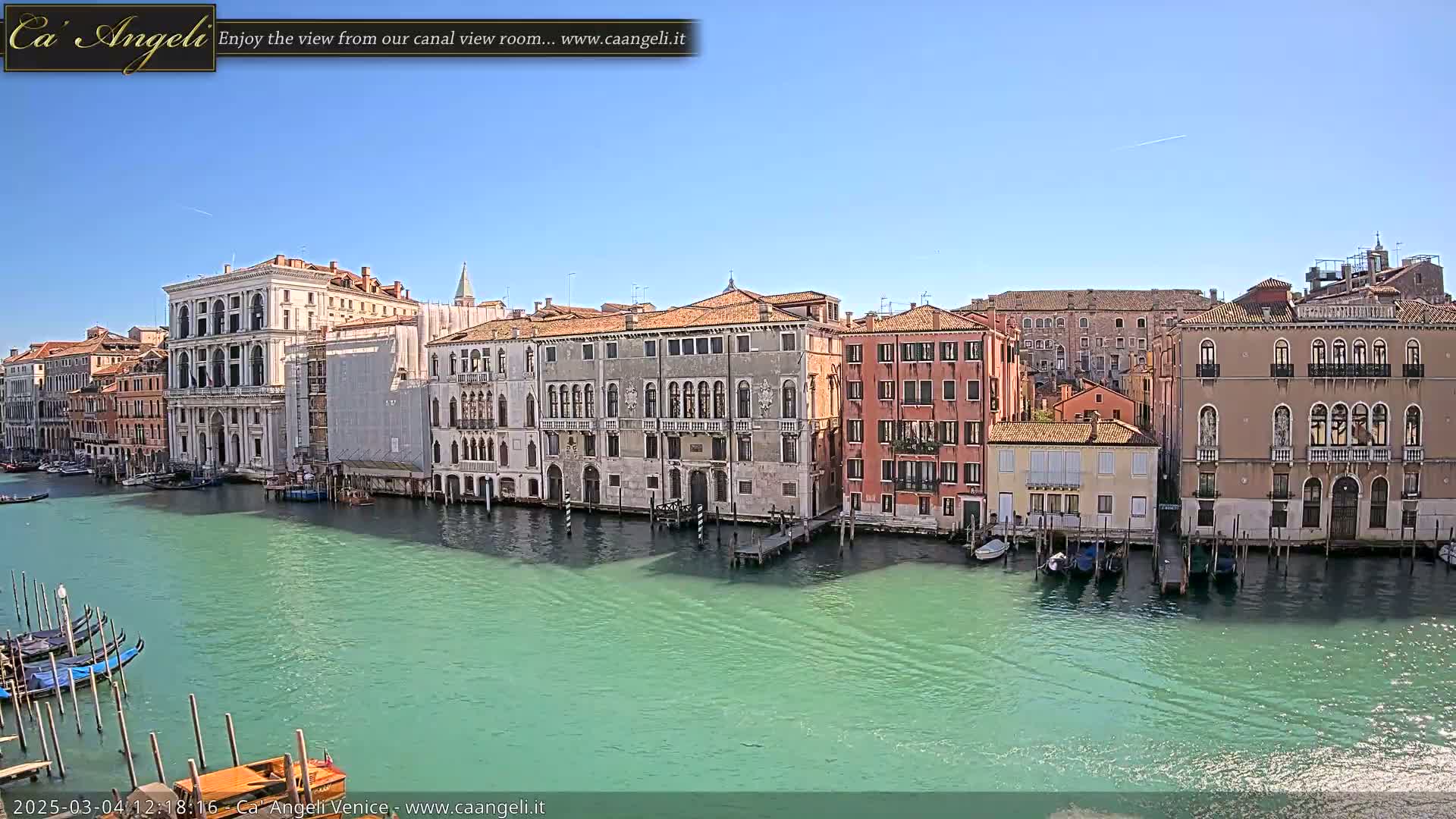 A picturesque canal in Venice, under a clear blue sky, is lined with historic buildings and several gondolas are moored along its edge.