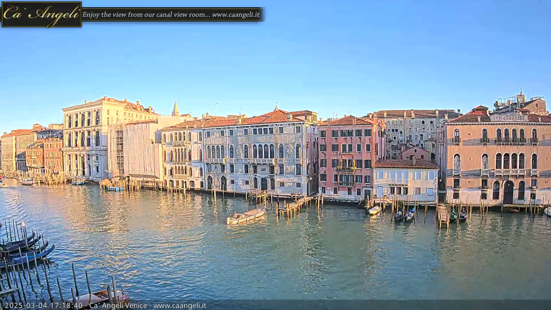 Under a clear blue sky, a canal in Venice is lined with several historic buildings, and gondolas are moored along its edges.
