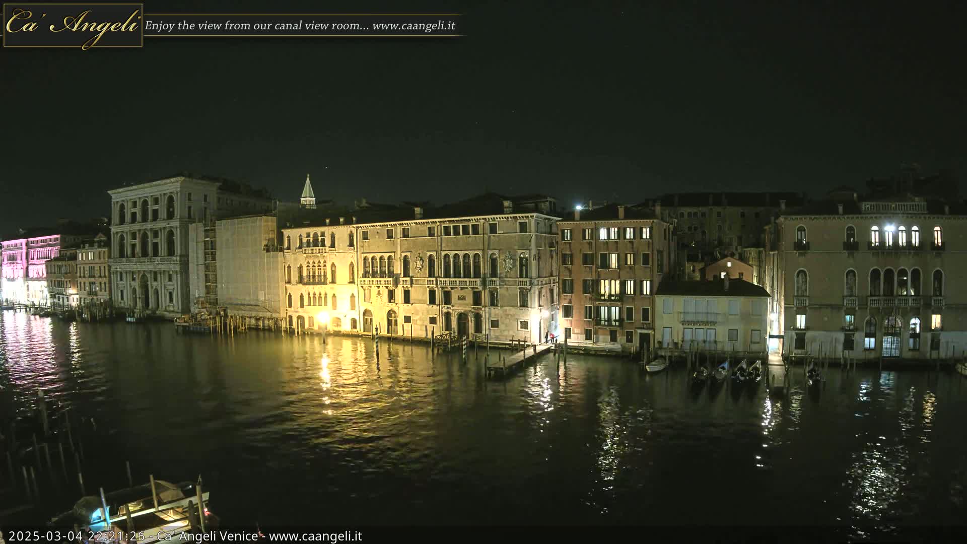 A nighttime view of a canal in Venice, Italy, shows several buildings illuminated along the waterway, with gondolas moored at the water's edge.
