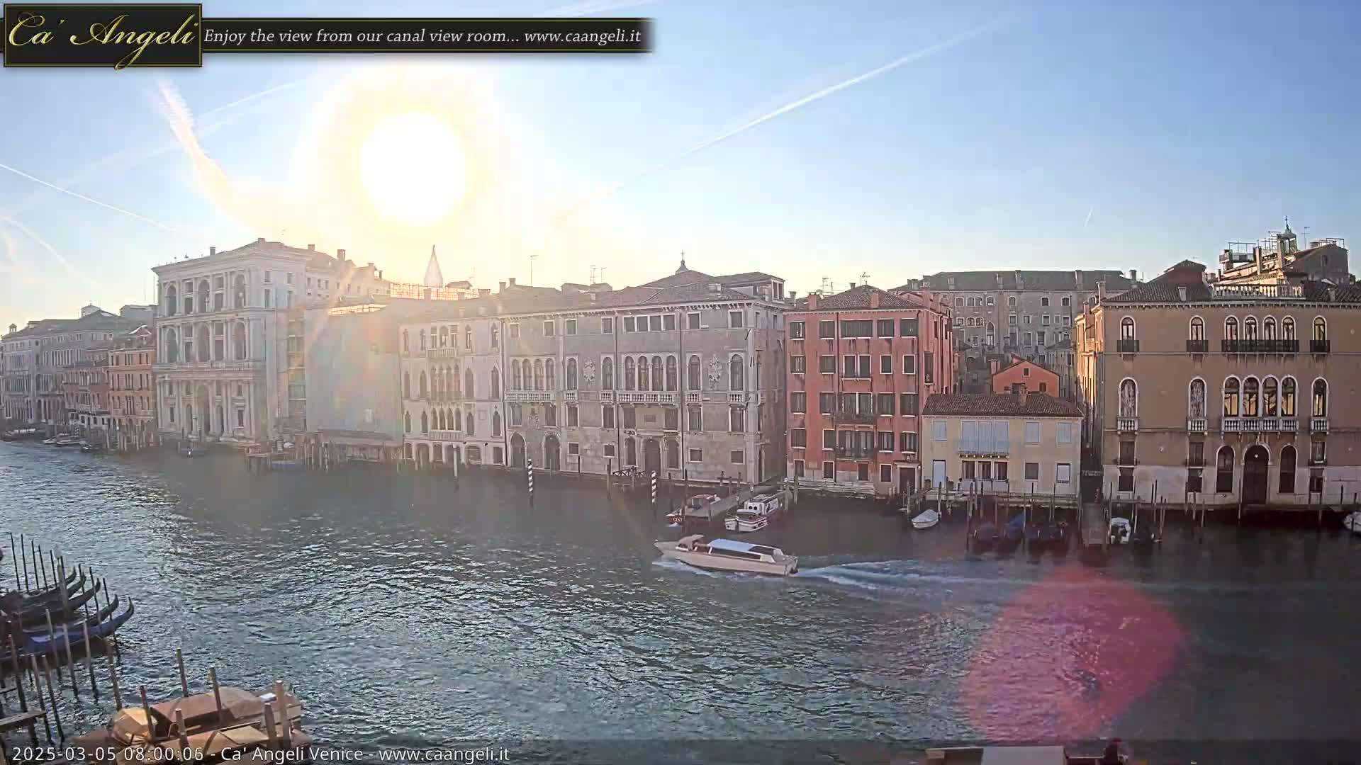 A bright, sunny day in Venice shows a canal lined with buildings, gondolas, and a motorboat.