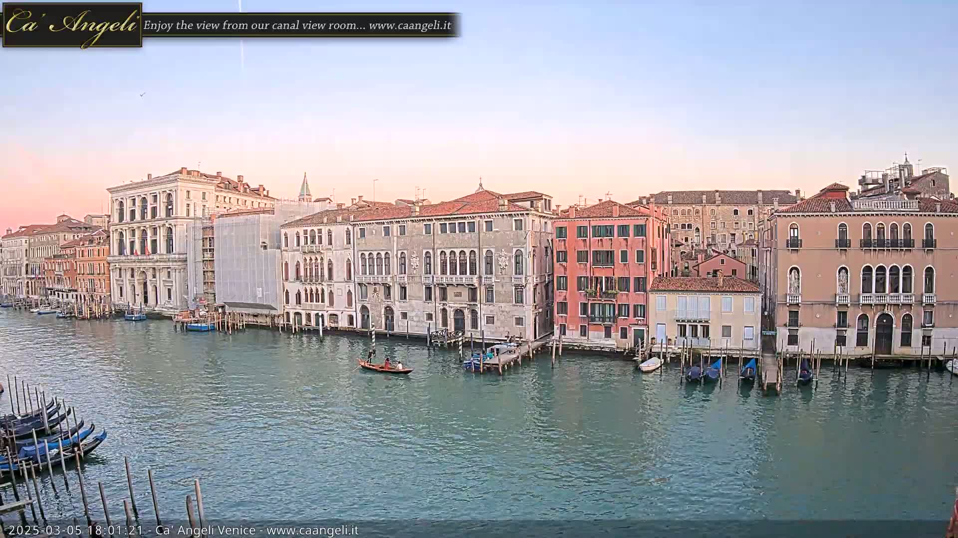 A gondola floats on a calm canal in Venice, past several historic buildings under a clear, pale sky at dusk.