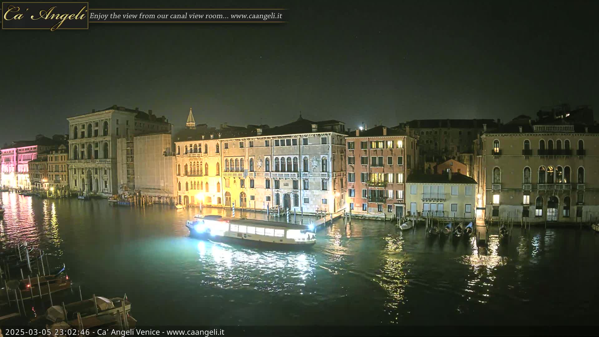 A nighttime view of a canal in Venice, Italy, shows several illuminated buildings along the waterway, with a boat traveling down the canal.