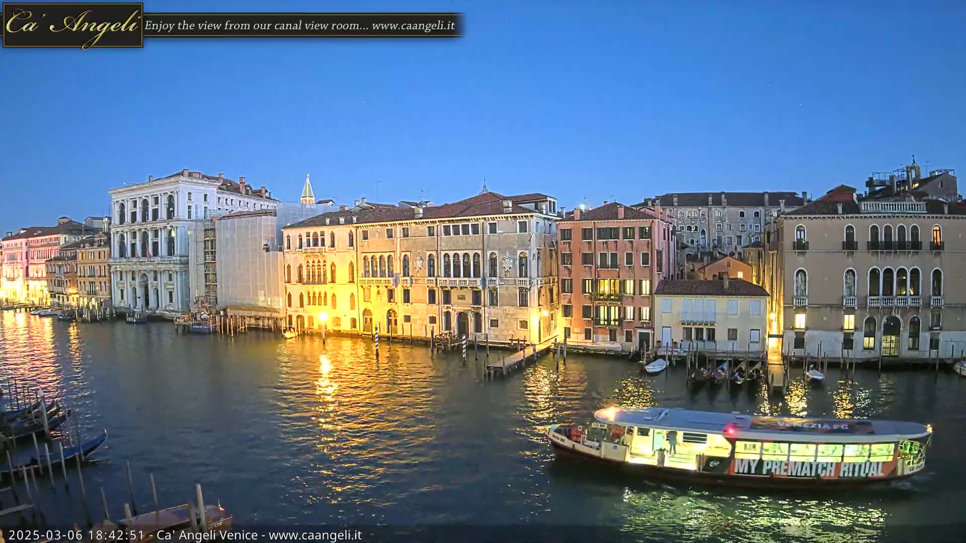 A water taxi travels along a canal at dusk, passing illuminated buildings in Venice under a clear blue sky.
