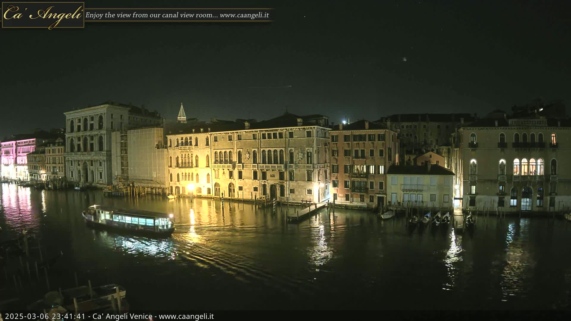 A nighttime view of a canal in Venice, Italy, shows several illuminated buildings lining the waterway, with a boat traveling down the canal, under a clear, starry sky.